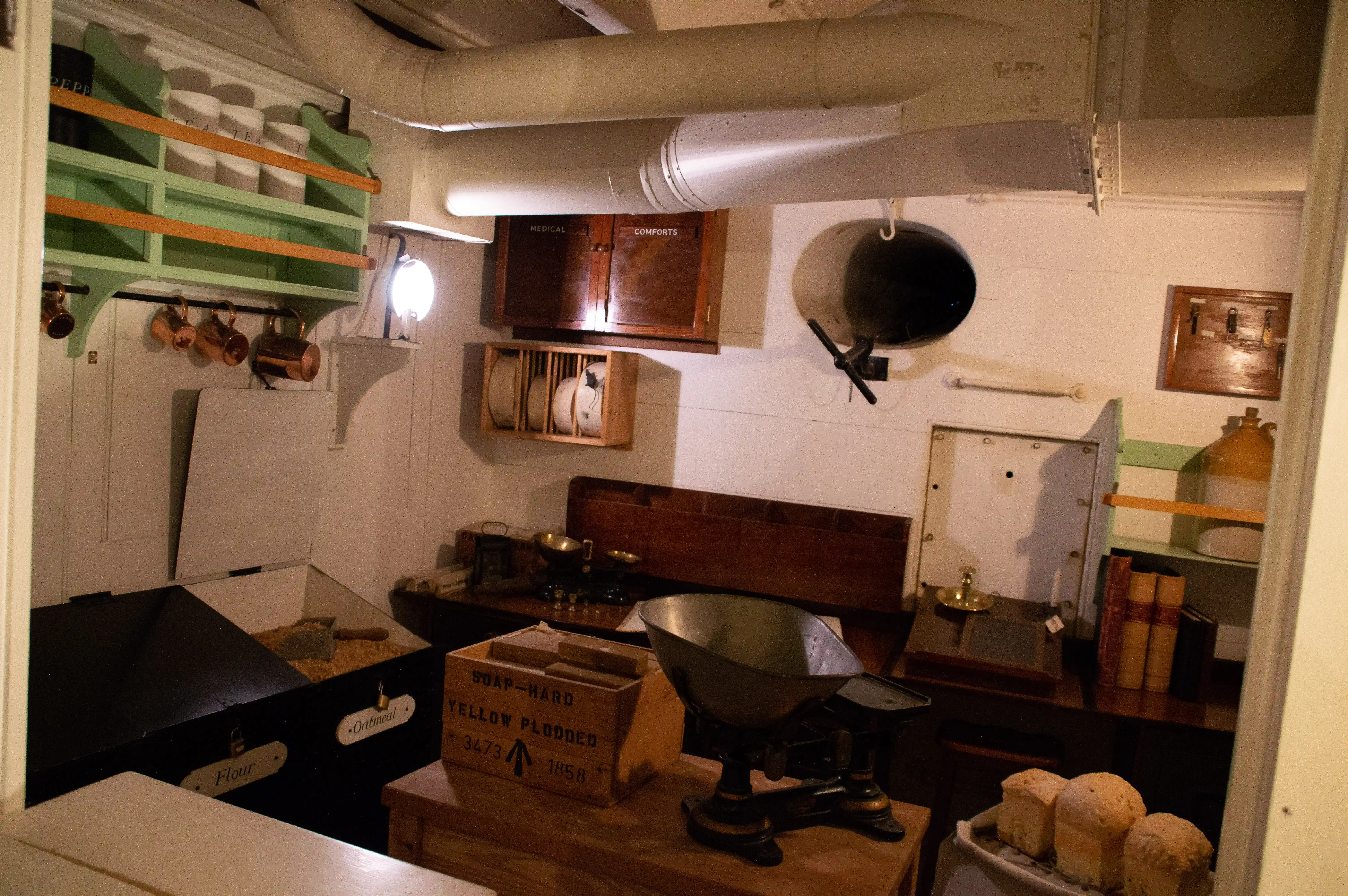The image depicts a historical ship's galley or kitchen area, featuring various food storage and preparation items. There are labeled containers for flour, oatmeal, and soap, a large wooden cabinet marked 'Medical' and 'Comforts', copper pots hanging on the wall, a weighing scale with a large metal bowl, and shelves stocked with supplies. The setting is compact and utilitarian, reflecting the limited space available in a ship's kitchen.