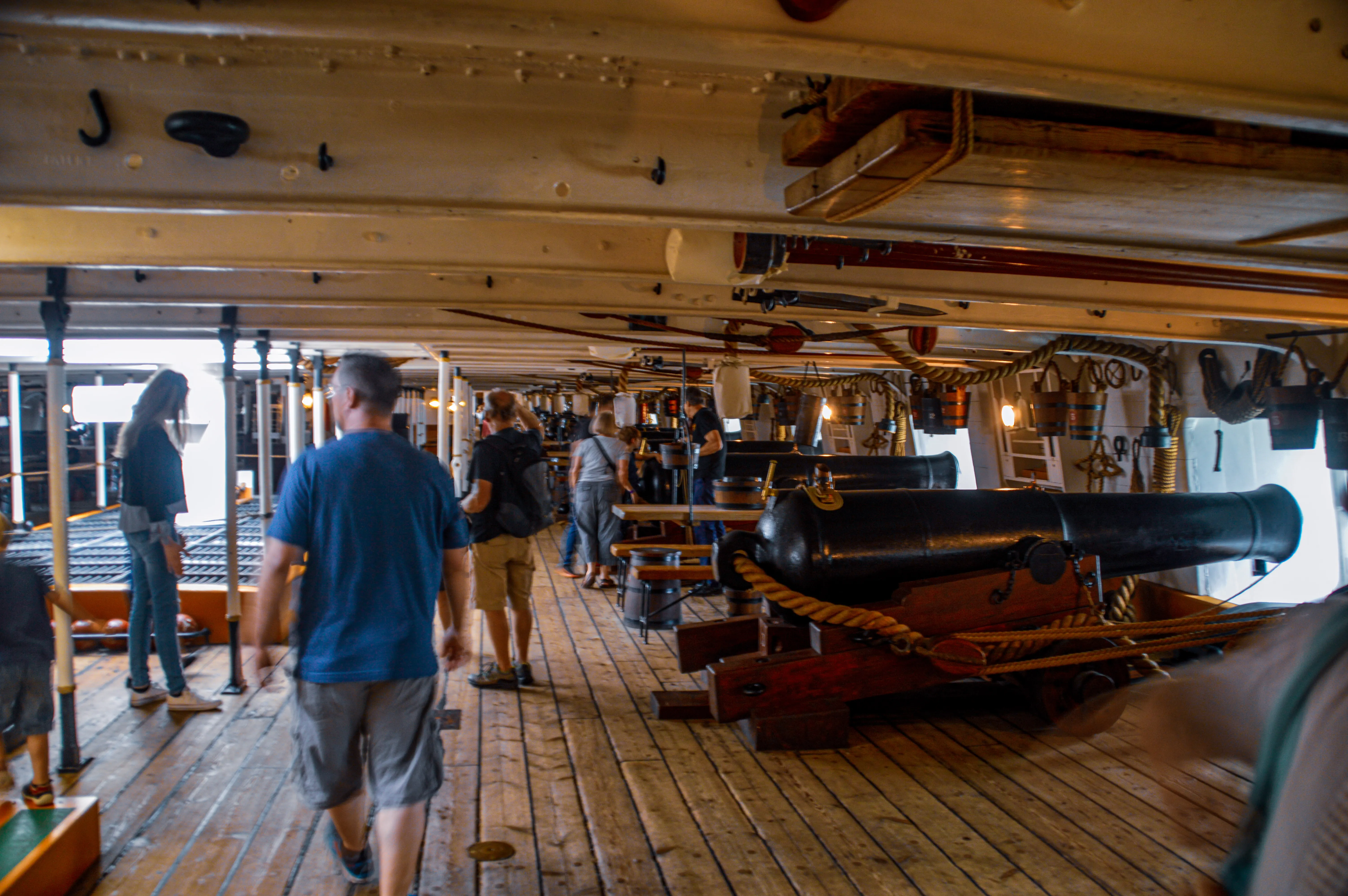 The image depicts the interior deck of an old wooden ship, likely a historical or replica vessel, with several people exploring it. The deck features wooden planks and beams, and is equipped with cannons and various nautical equipment. The visitors appear to be tourists, observing the ship's features and structure.