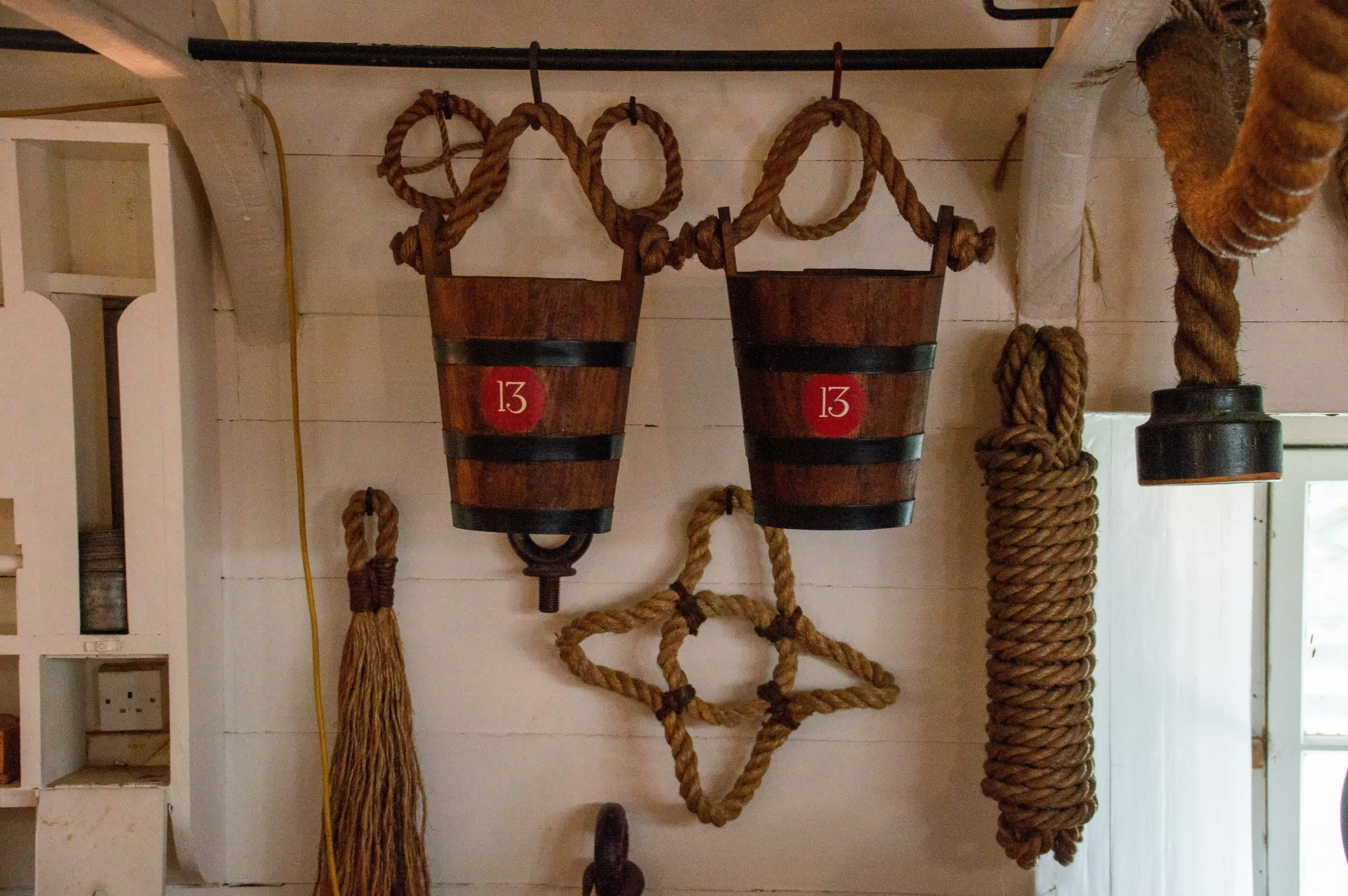 The image shows a nautical-themed display with two wooden buckets labeled '13' hanging from ropes, a coiled rope star, a large rope coil, a broom, and other maritime items arranged on a white wooden wall.