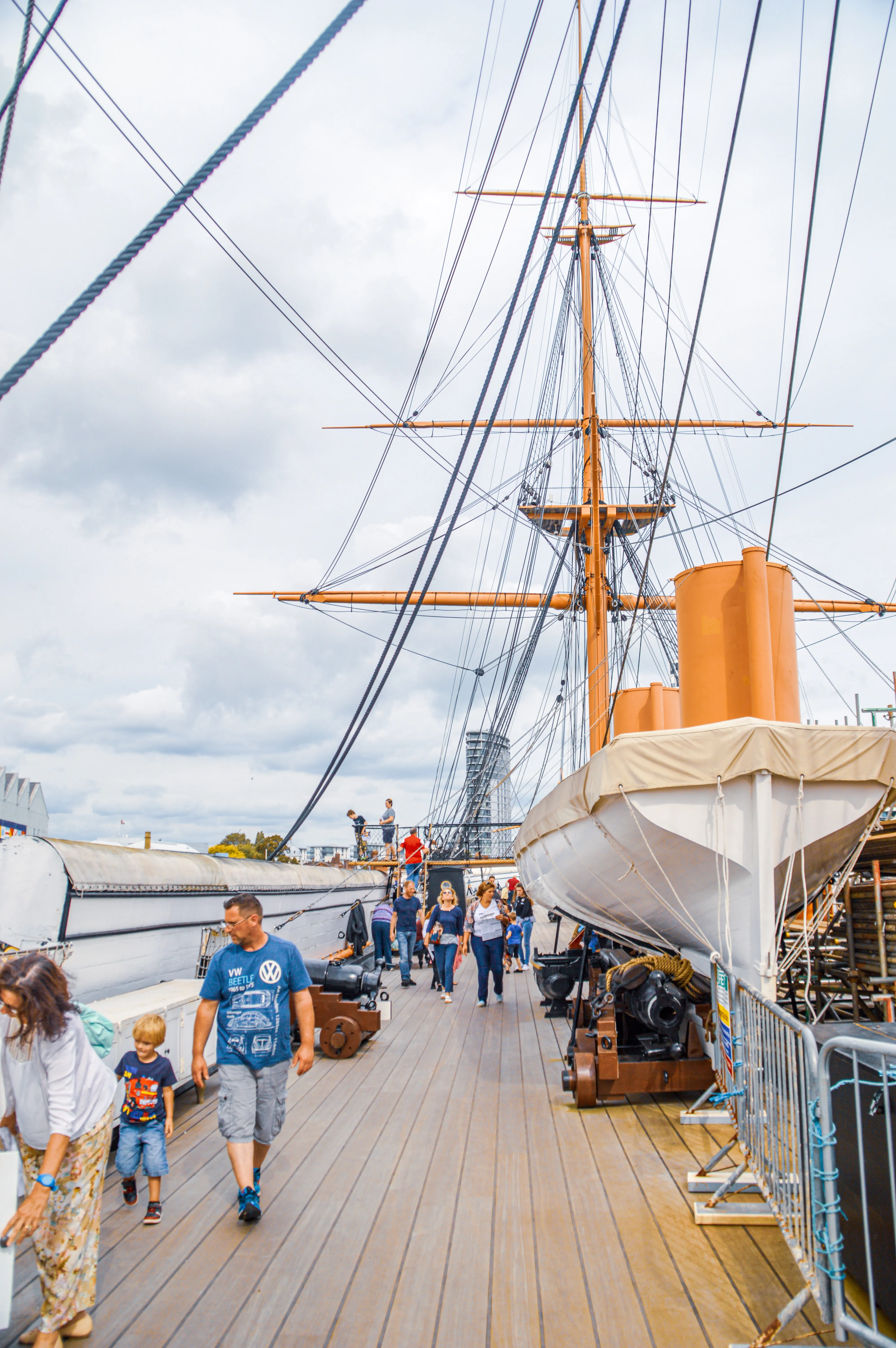 This image captures a lively scene on the wooden deck of what appears to be a historic sailing ship. A number of people are strolling along the deck, taking in the sights. Dominating the background is the ship's impressive mast and rigging, a testament to its grand past. In the foreground, a life-saving boat is neatly stowed, and a couple of old cannons add to the nautical atmosphere. A modern city skyline is visible in the distance, creating a fascinating contrast between old and new