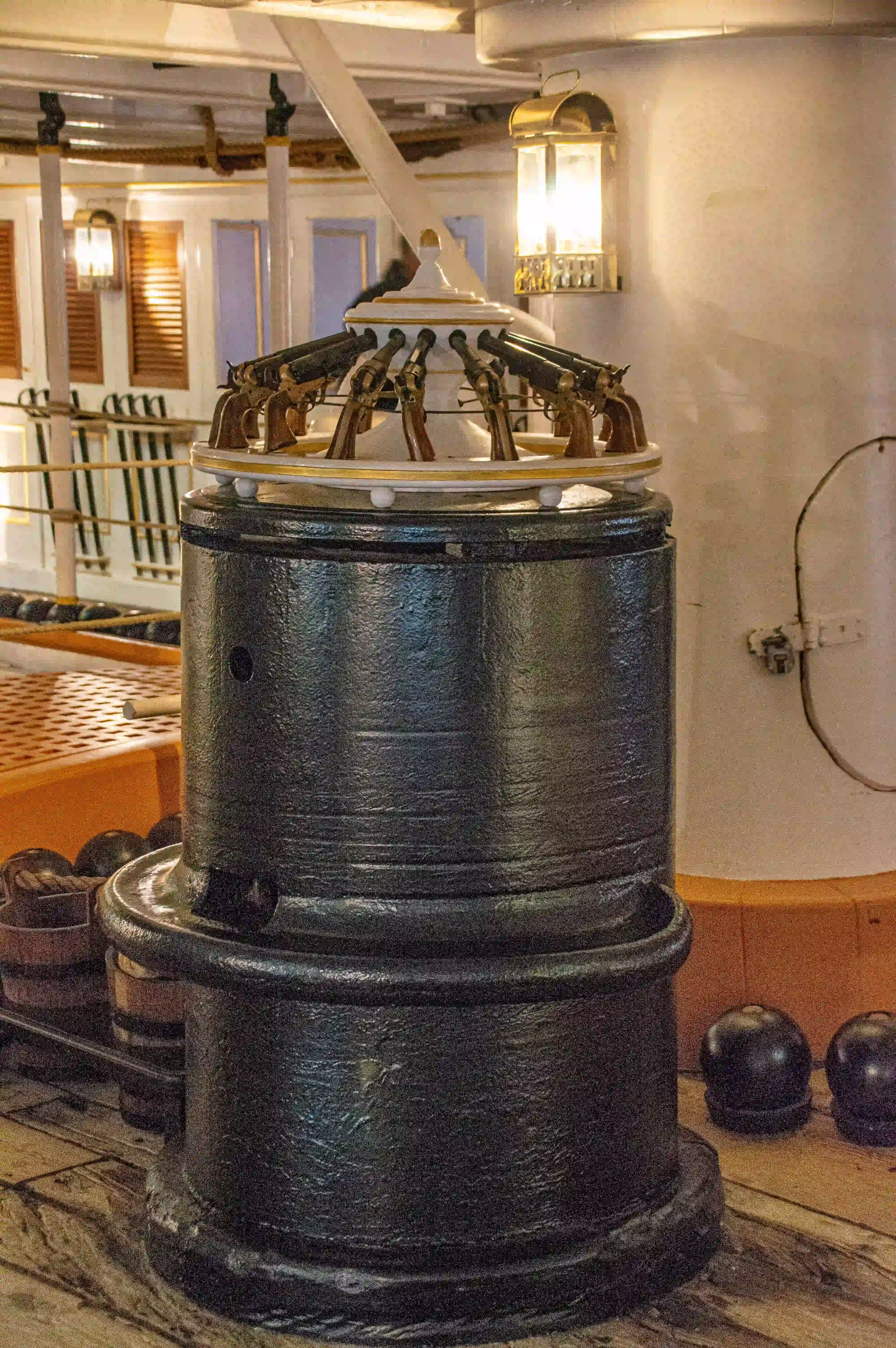 The image depicts an old naval ship cannon mounted on a wooden deck. The cannon is a multi-barreled design, likely a revolver cannon, with multiple barrels arranged in a circular fashion around a central axis. The setting appears to be an indoor display, possibly in a museum or a historical ship replica, with wooden beams, lanterns, and other maritime elements visible in the background.