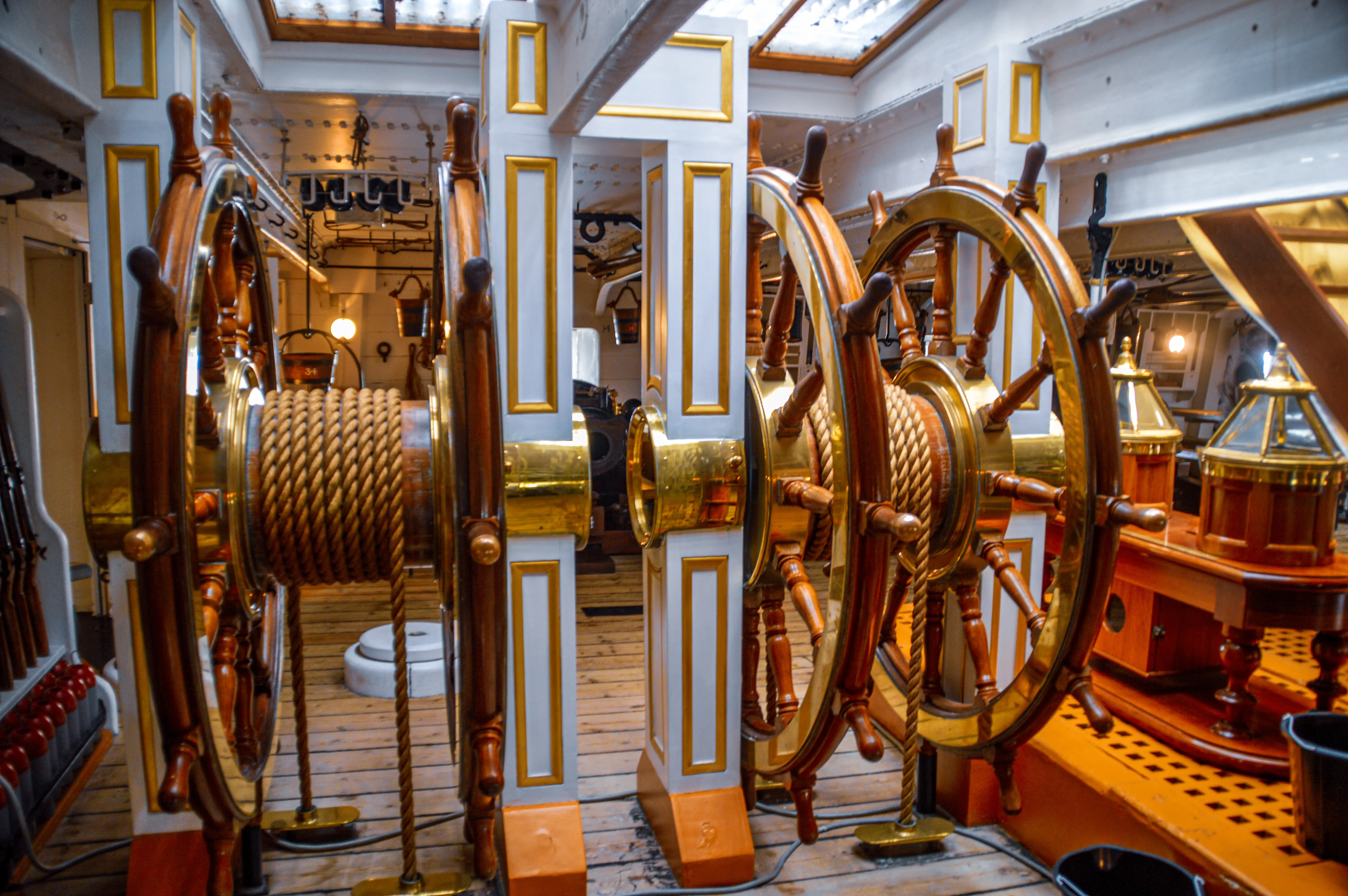 The image depicts the interior of a ship, showcasing ornate steering wheels and ropes, with a well-maintained and polished wooden deck and walls. The area appears to be part of the ship's control or navigation section, featuring intricate gold and wood detailing. The setting suggests a historical or museum context, emphasizing the ship's vintage design and craftsmanship.