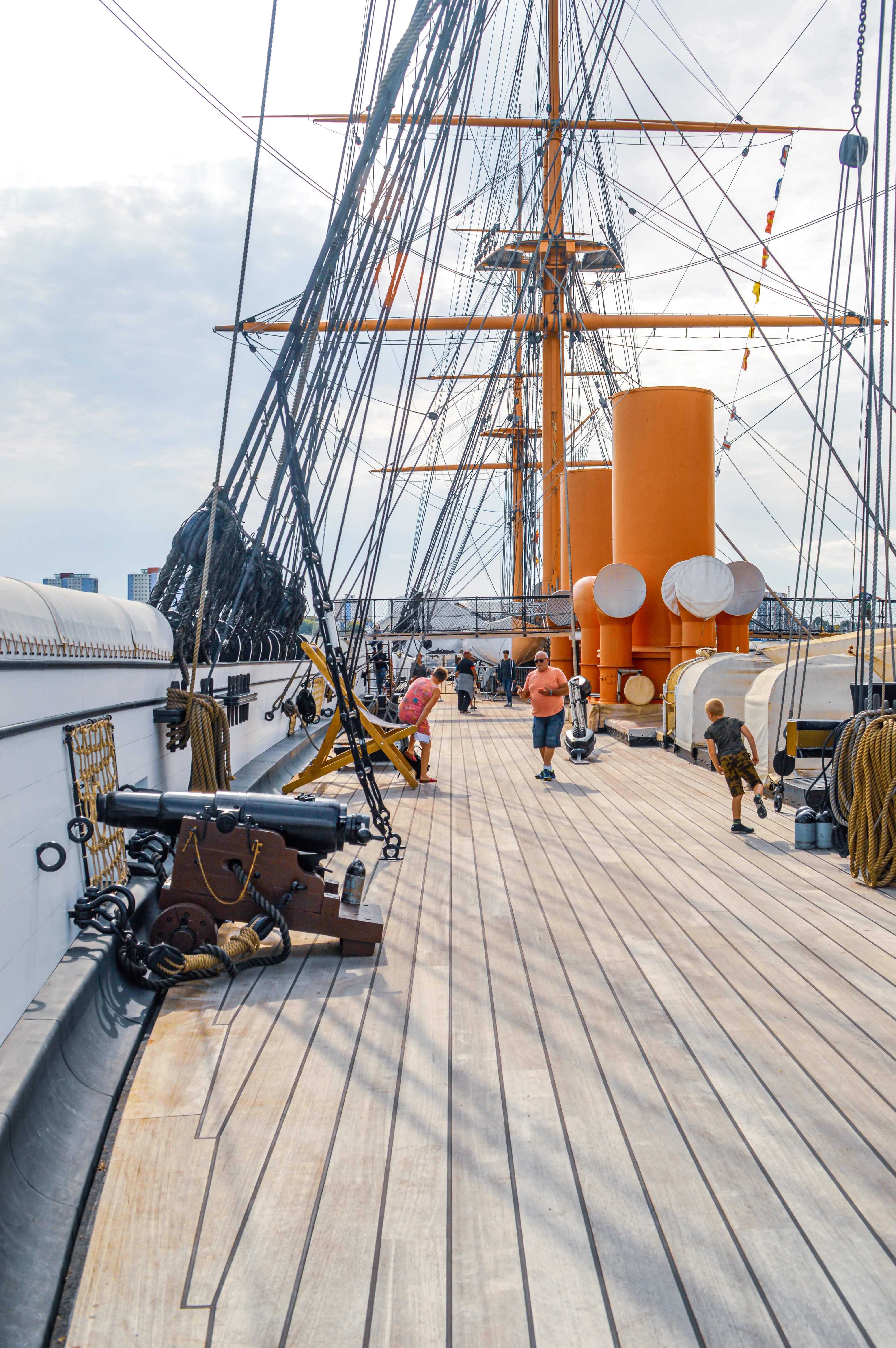 The image shows a lovely view from the deck of a historic sailing ship, bathed in bright sunshine. We can see a wooden deck with several people enjoying the day, including children. In the foreground, there's an old-fashioned cannon, adding to the maritime atmosphere. The masts and rigging of the ship stretch up towards a slightly cloudy sky, giving us a sense of its grandeur. The overall impression is one of a pleasant and engaging visit to a fascinating piece of naval history!