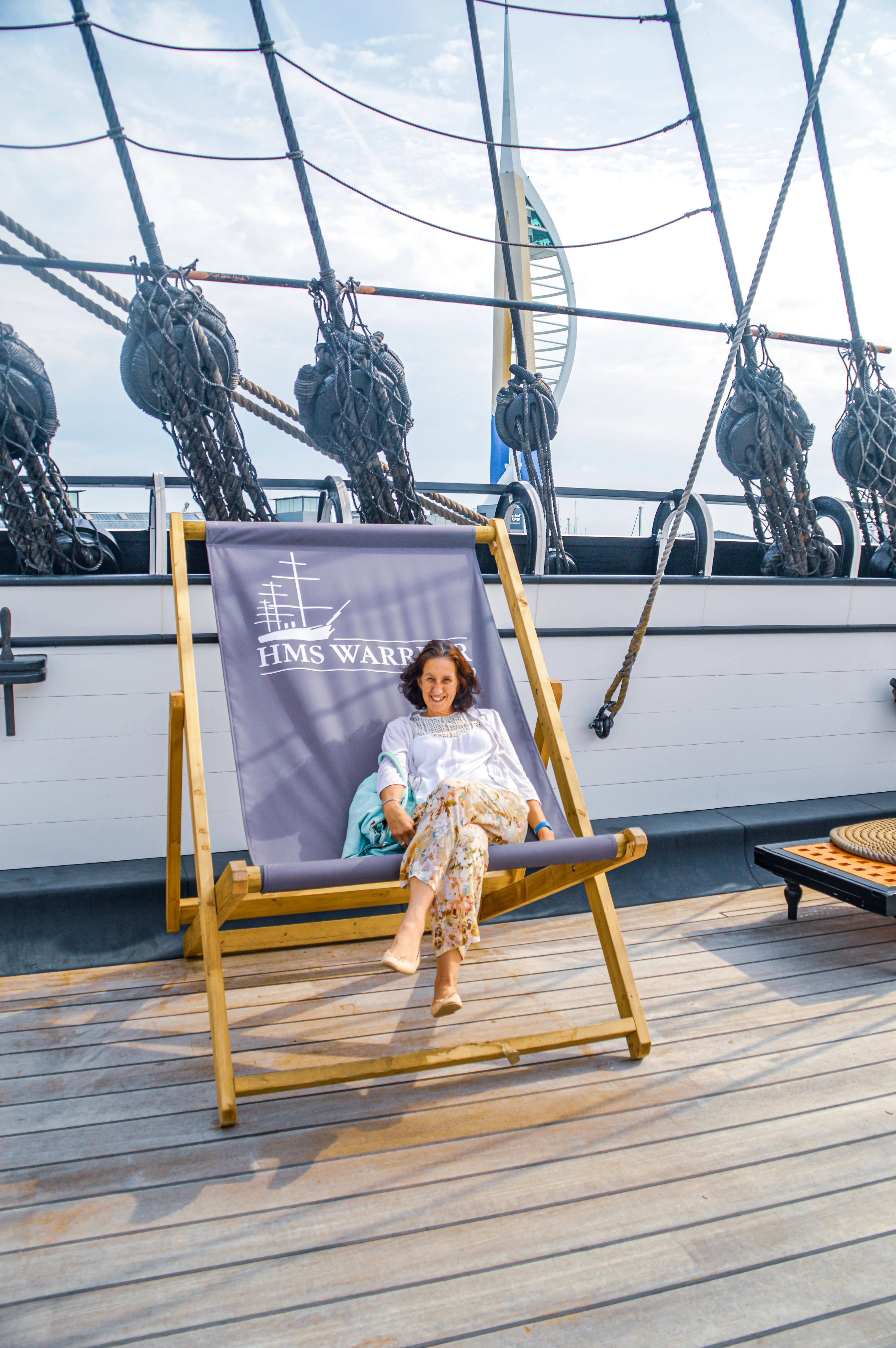 The image shows a person sitting on a deck chair on the deck of a ship named HMS Warrior. The ship has a lot of ropes and pulleys visible in the background, and the deck is made of wooden planks. The person is smiling and appears to be enjoying the sunny weather. In the background, part of a Ferris wheel can be seen, indicating that the ship might be docked near an amusement area.