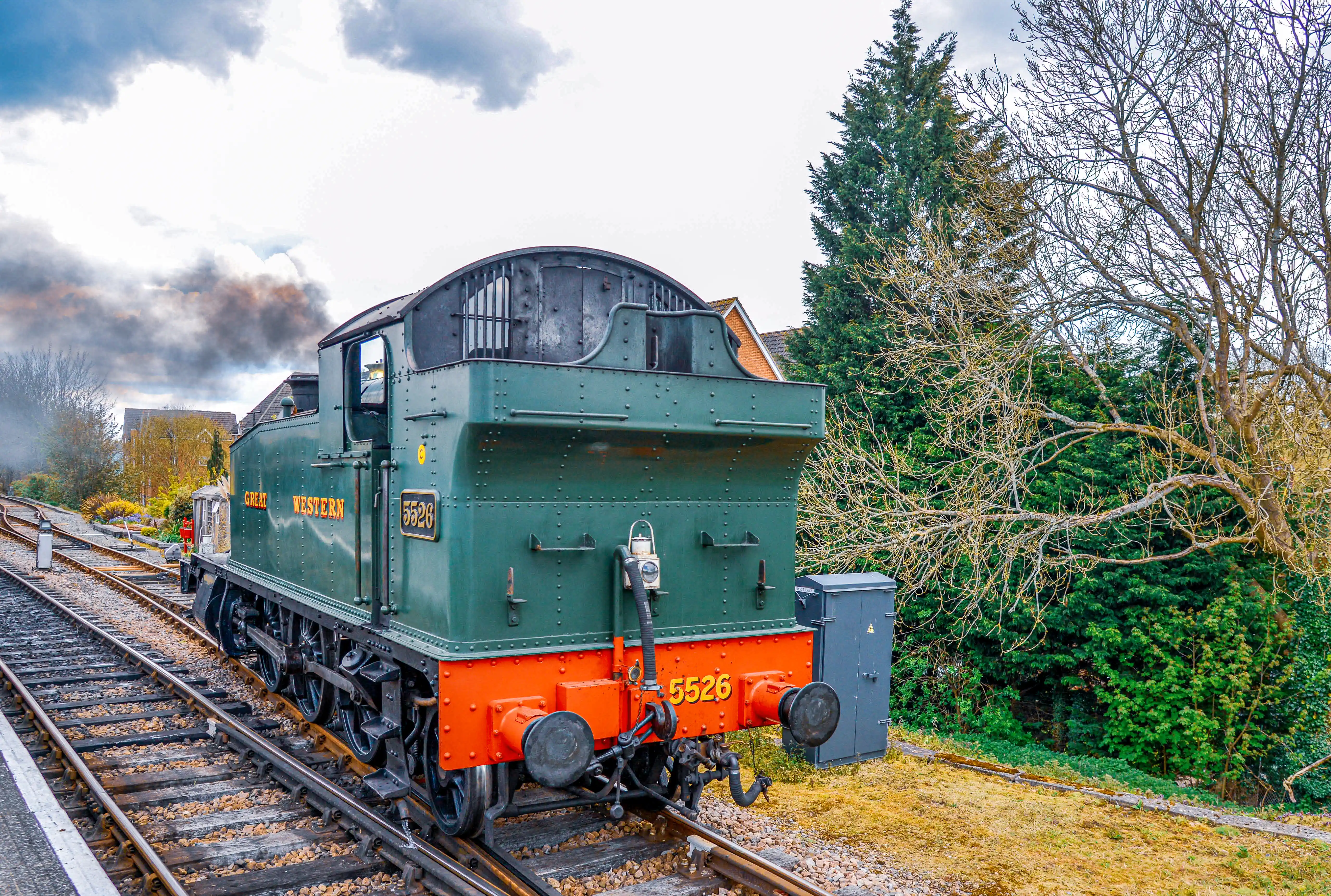 The image depicts a green steam locomotive with the number 5526 prominently displayed. The locomotive is on a railway track with some smoke visible, suggesting it is in operation or has recently been running. The surrounding area includes trees, a residential building, and a partly cloudy sky, indicating a serene, possibly rural setting.