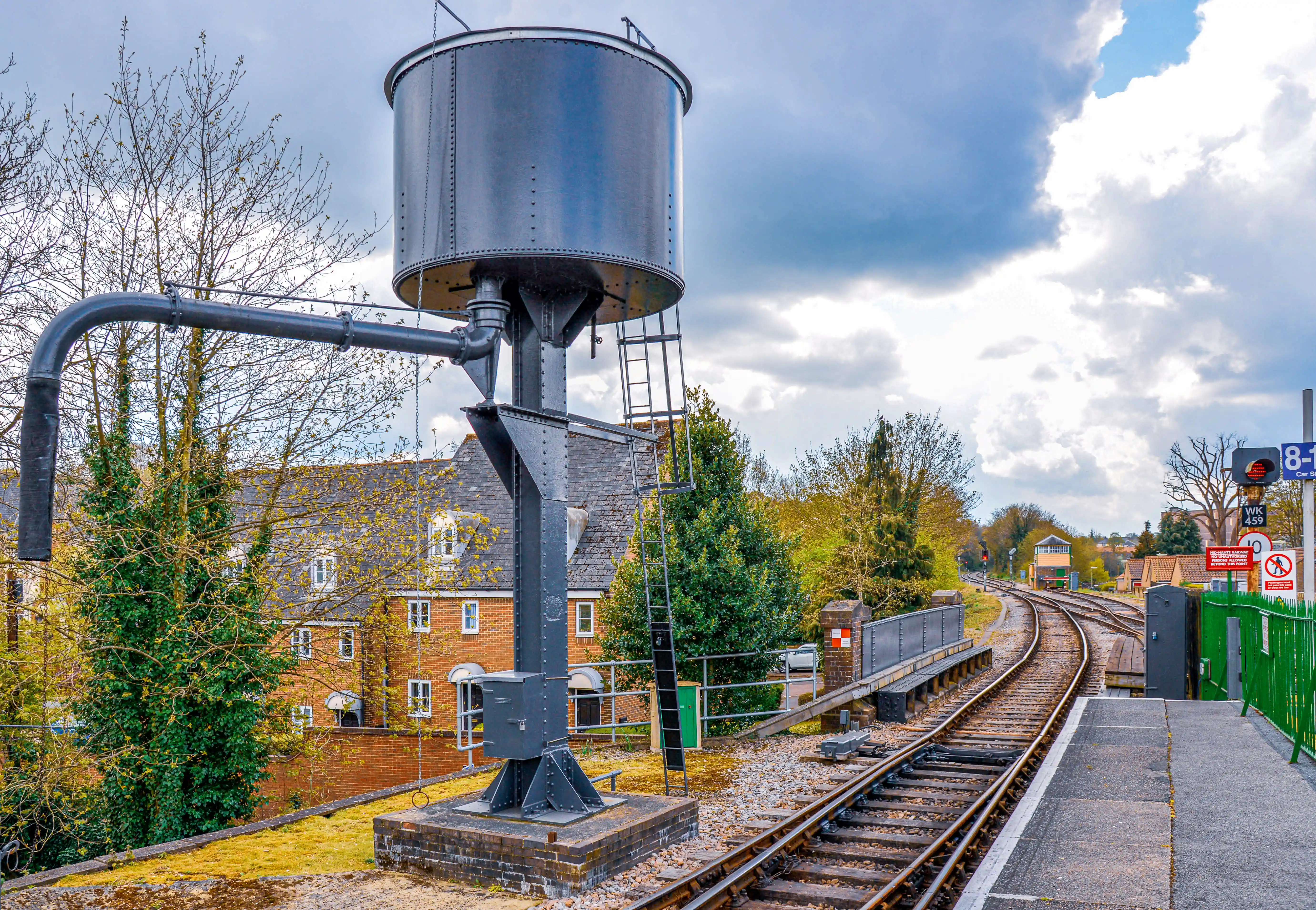 The image depicts a railway station with a prominent water tower structure adjacent to the tracks. The station platform is visible on the right, with railway tracks curving gently to the left. There are residential buildings in the background, along with some greenery and trees. The sky is partly cloudy, and the overall scene suggests a peaceful, suburban setting.