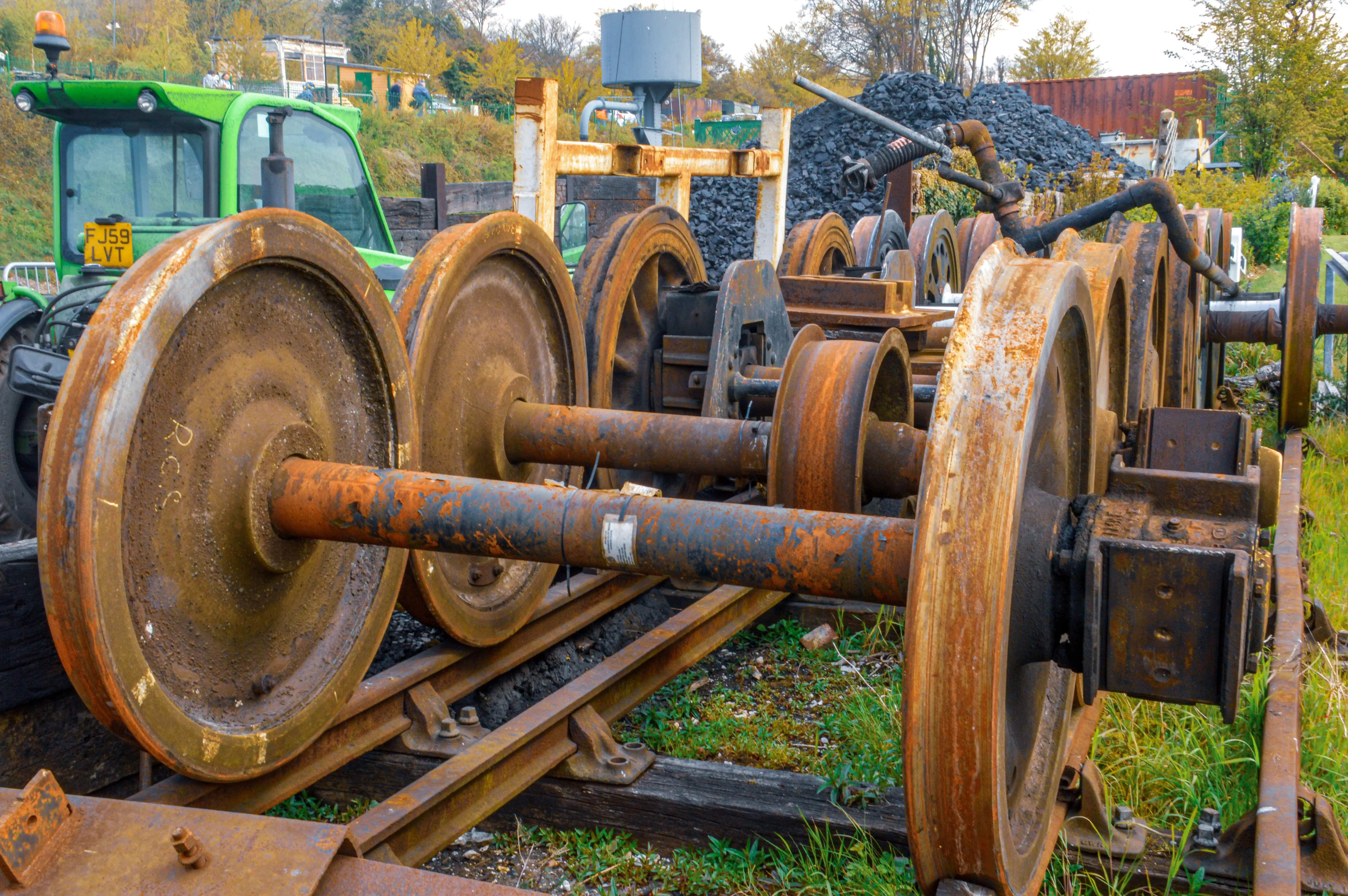 The image depicts a set of rusty train wheels on a railway track, with a green tractor in the background. The wheels are part of an old or abandoned railway system, showing signs of wear and rust. There is a pile of coal or similar material nearby, suggesting an industrial setting. The scene is set in an outdoor environment with greenery and some buildings visible in the distance.