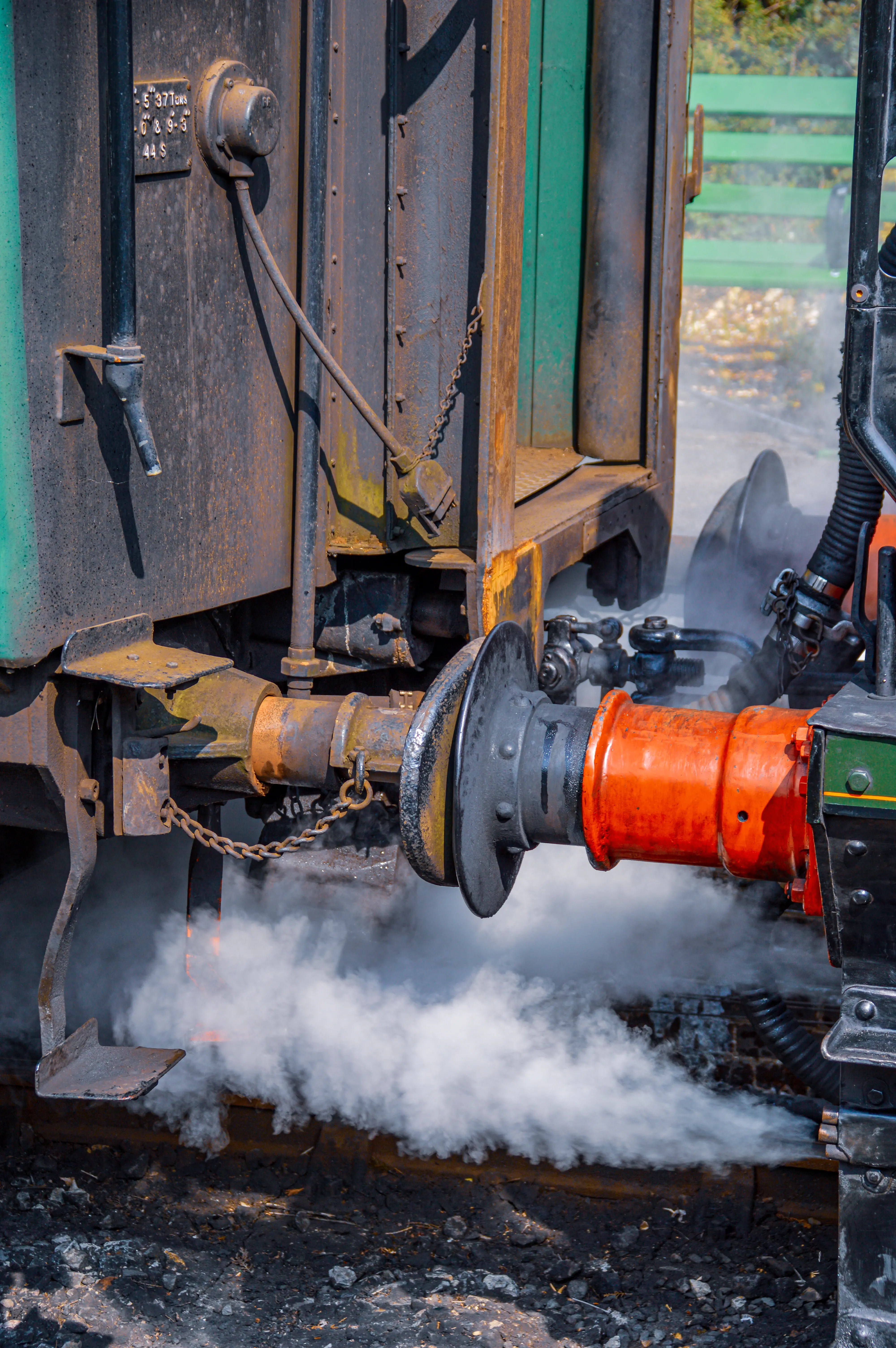 The image depicts a close-up view of a mechanical coupling between two train cars. Steam or smoke is visibly emanating from the connection area, suggesting recent detachment or attachment. The components shown include a chain, a cylindrical coupling mechanism, and various metal parts. The scene appears to be set on railway tracks with some greenery visible in the background.
