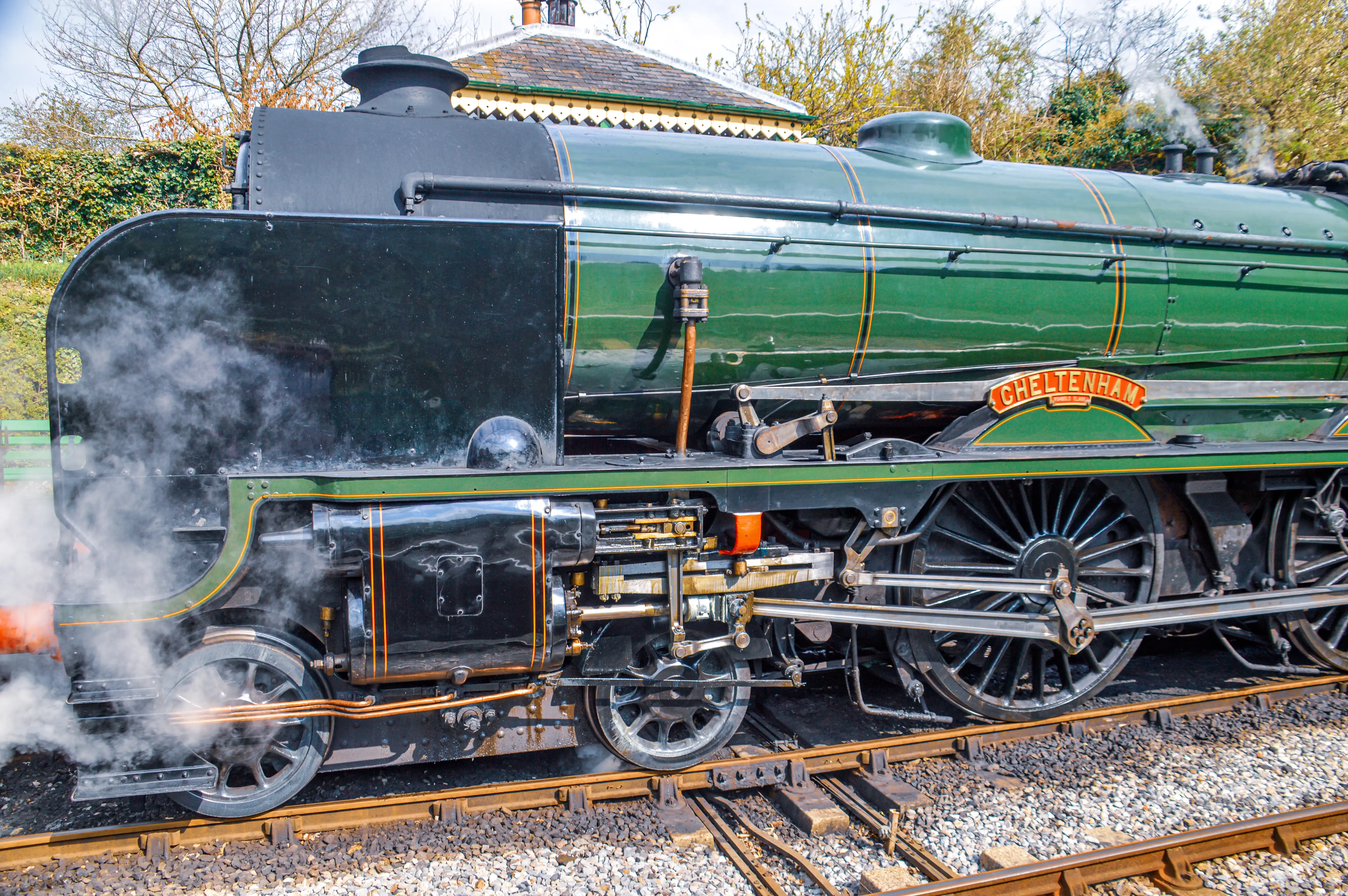 The image depicts a close-up view of a vintage steam locomotive named 'Cheltenham.' The locomotive is painted in a dark green color with gold accents. Steam can be seen emitting from the engine, indicating it is in operation. The intricate mechanical details of the wheels and pistons are prominently visible.