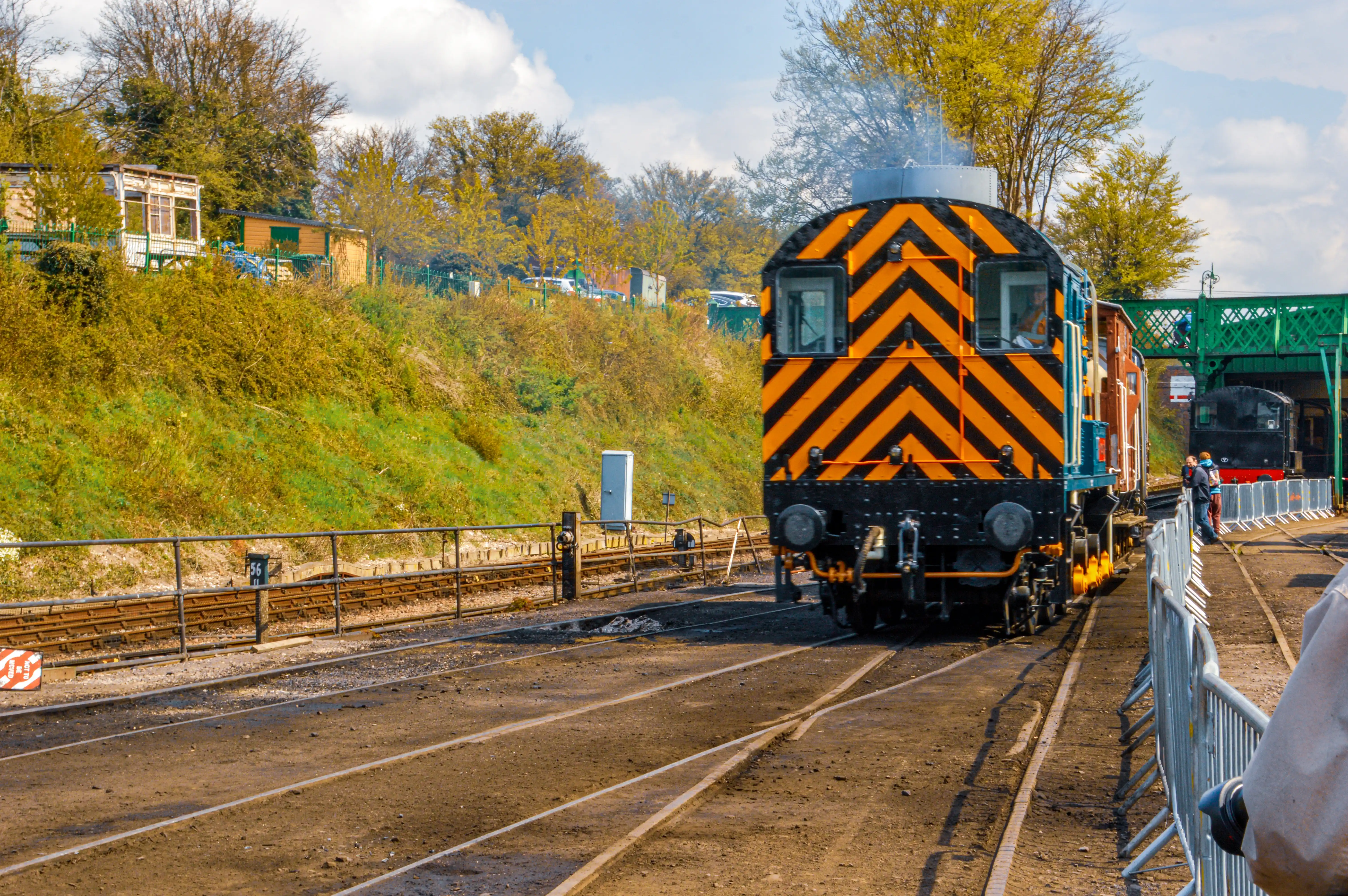 The image depicts a railway scene with a prominently displayed train engine painted in orange and black diagonal stripes. The train is situated on the tracks, with some greenery and buildings visible in the background. There are a few people visible near the train and tracks, and the setting appears to be a railway station or yard. The weather seems clear with some clouds in the sky.
