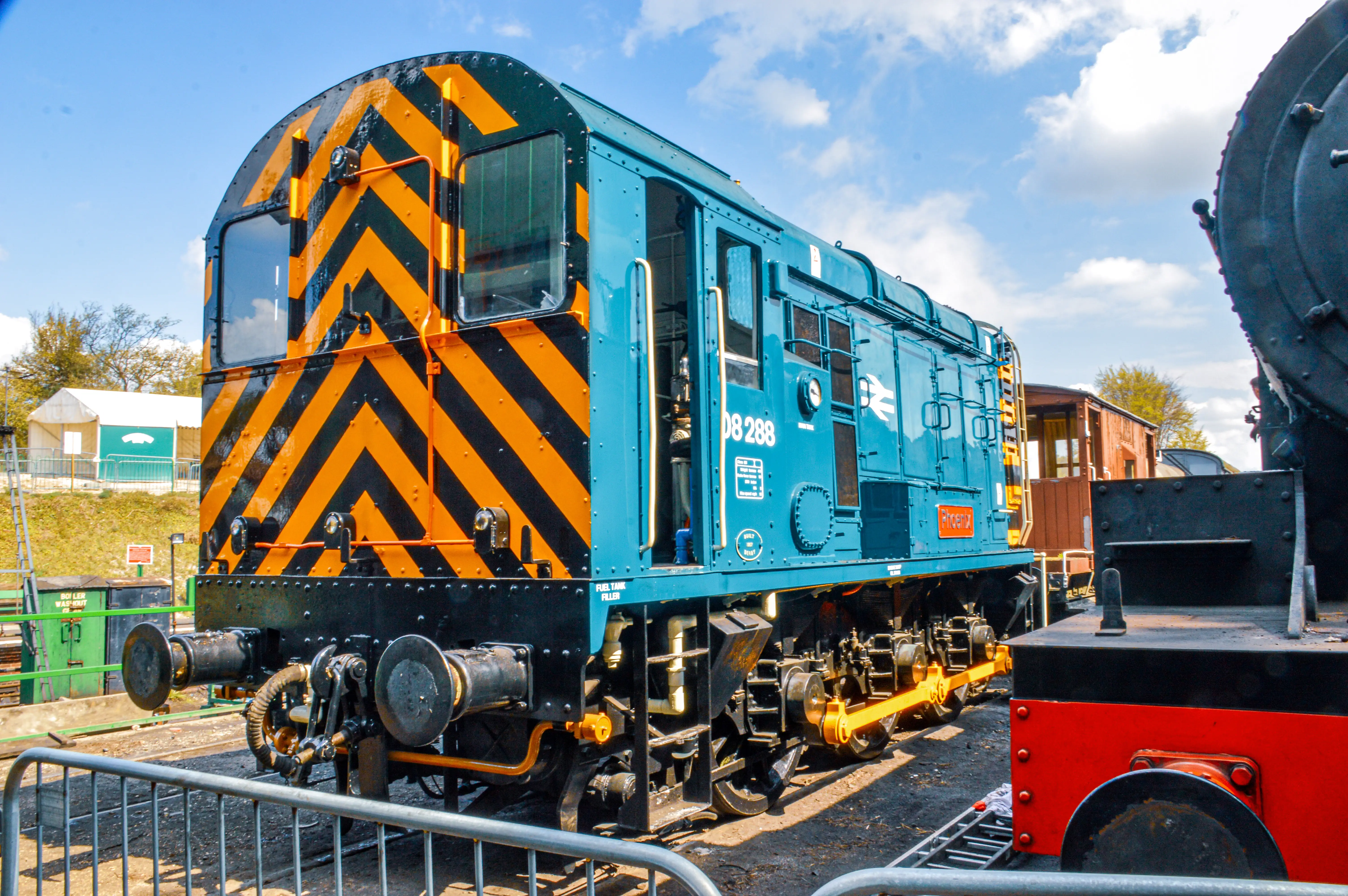 The image depicts a blue and yellow train engine with black diagonal stripes, labeled with the number 08 288. The train is situated on tracks in an outdoor setting, possibly a railway museum or a train yard. The environment includes some greenery and buildings in the background, with another train partially visible on the right side of the image.