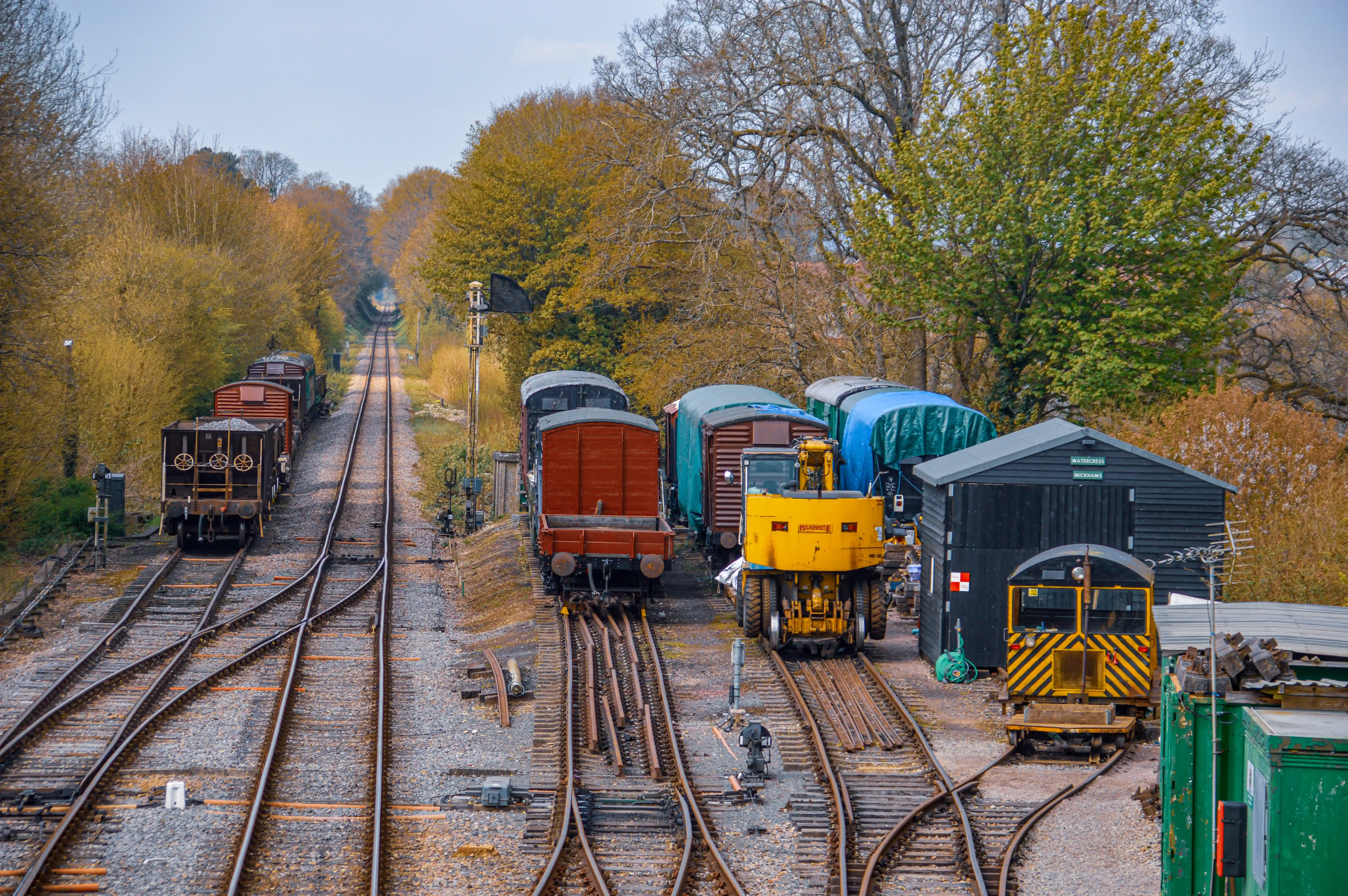 The image depicts a railway yard with multiple tracks converging and diverging. Several railcars and maintenance vehicles are stationed on the tracks. The scene is set in a wooded area with autumn foliage, and there are small buildings and sheds alongside the tracks. The overall atmosphere is calm and industrial, with a focus on railway operations and maintenance.