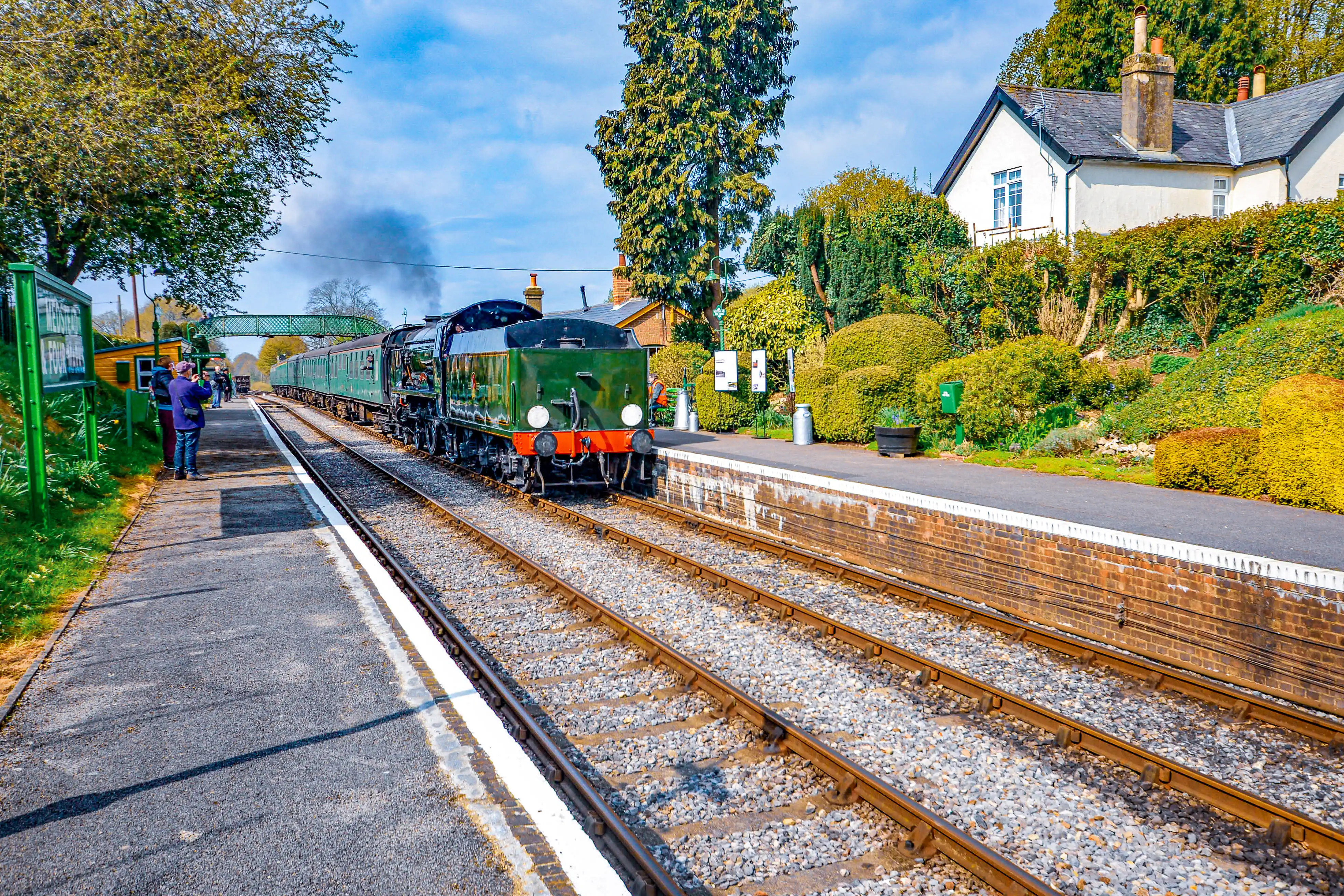 The image depicts a vintage steam train arriving at a quaint, well-maintained railway station. The station platform is adorned with greenery and has a few people waiting. The train is green with a black and red front, and it is pulling several passenger carriages. The surrounding area features houses and lush vegetation, indicating a picturesque, possibly rural setting.