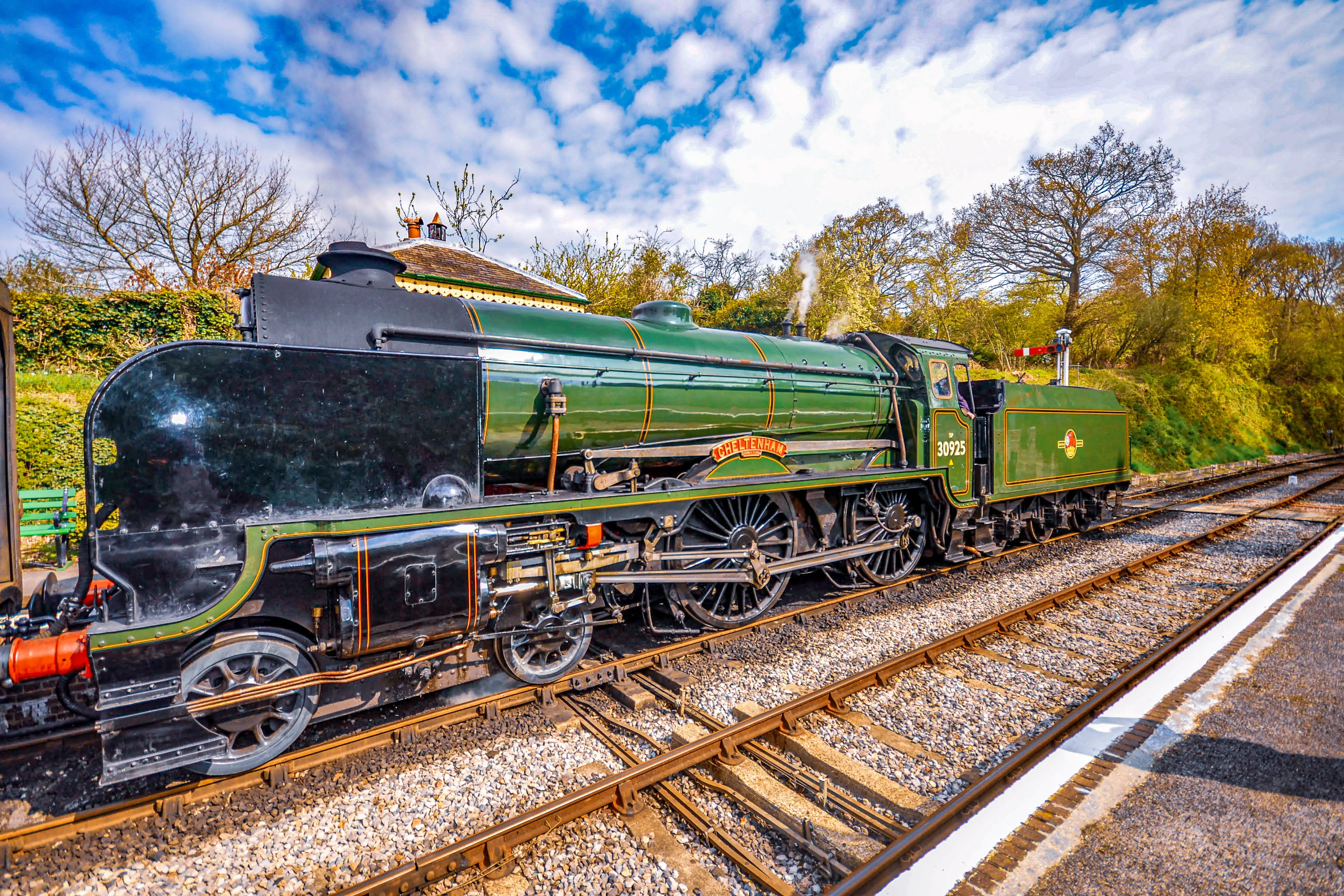 The image depicts a vintage steam locomotive, painted primarily in green with black and gold accents, positioned on railway tracks. The train is situated in a picturesque outdoor setting with trees and a partly cloudy sky in the background. The locomotive appears to be well-maintained, showcasing intricate details and craftsmanship typical of historic trains. The scene suggests a heritage railway setting, likely aimed at preserving and showcasing historical trains for educational or recreational purposes.