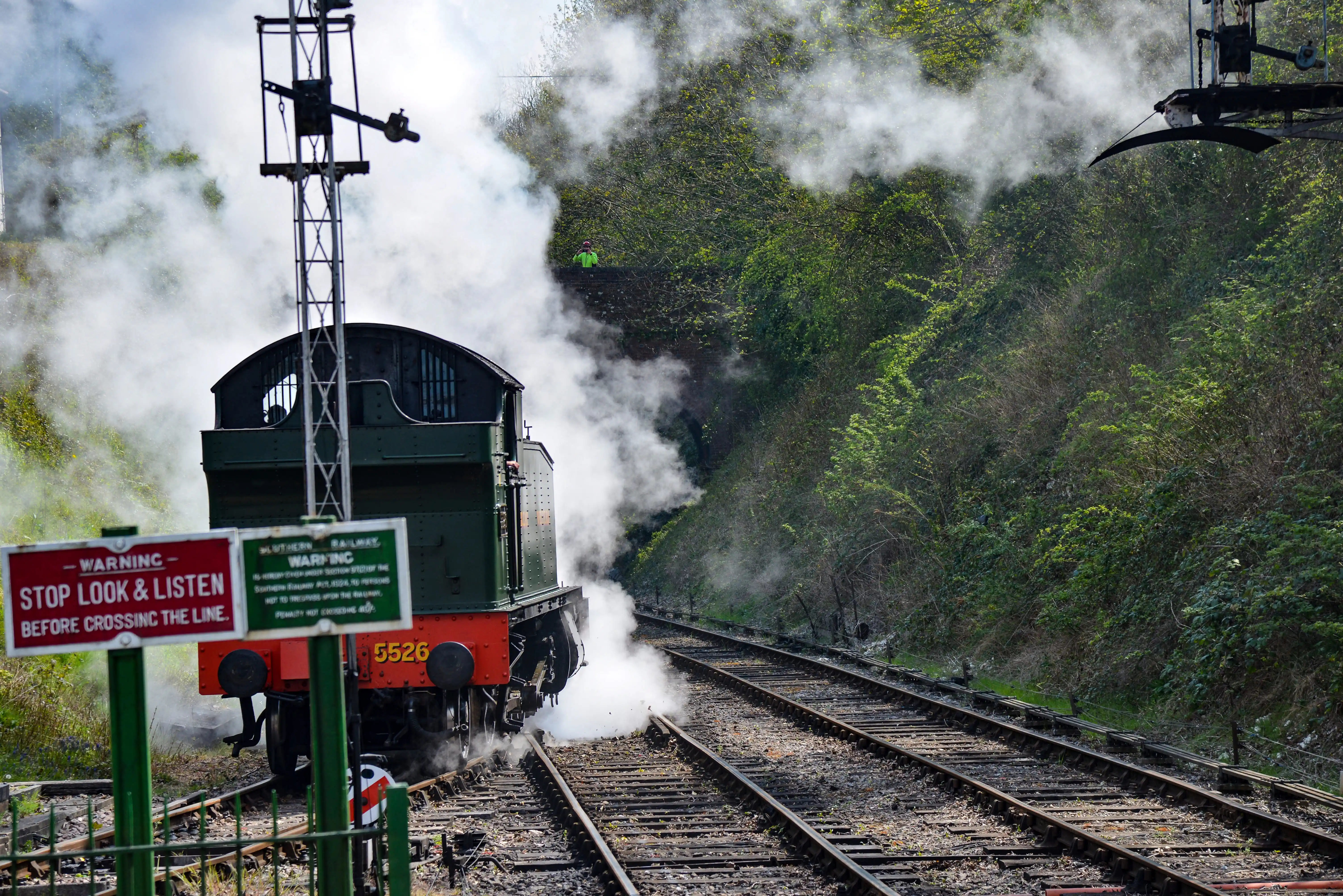 The image depicts a steam locomotive emitting steam as it travels along railway tracks. There is a warning sign in the foreground that reads 'STOP LOOK & LISTEN BEFORE CROSSING THE LINE.' The locomotive is green with red accents and has the number 5526 visible. The scene is set in a lush, green environment with dense foliage on either side of the tracks.