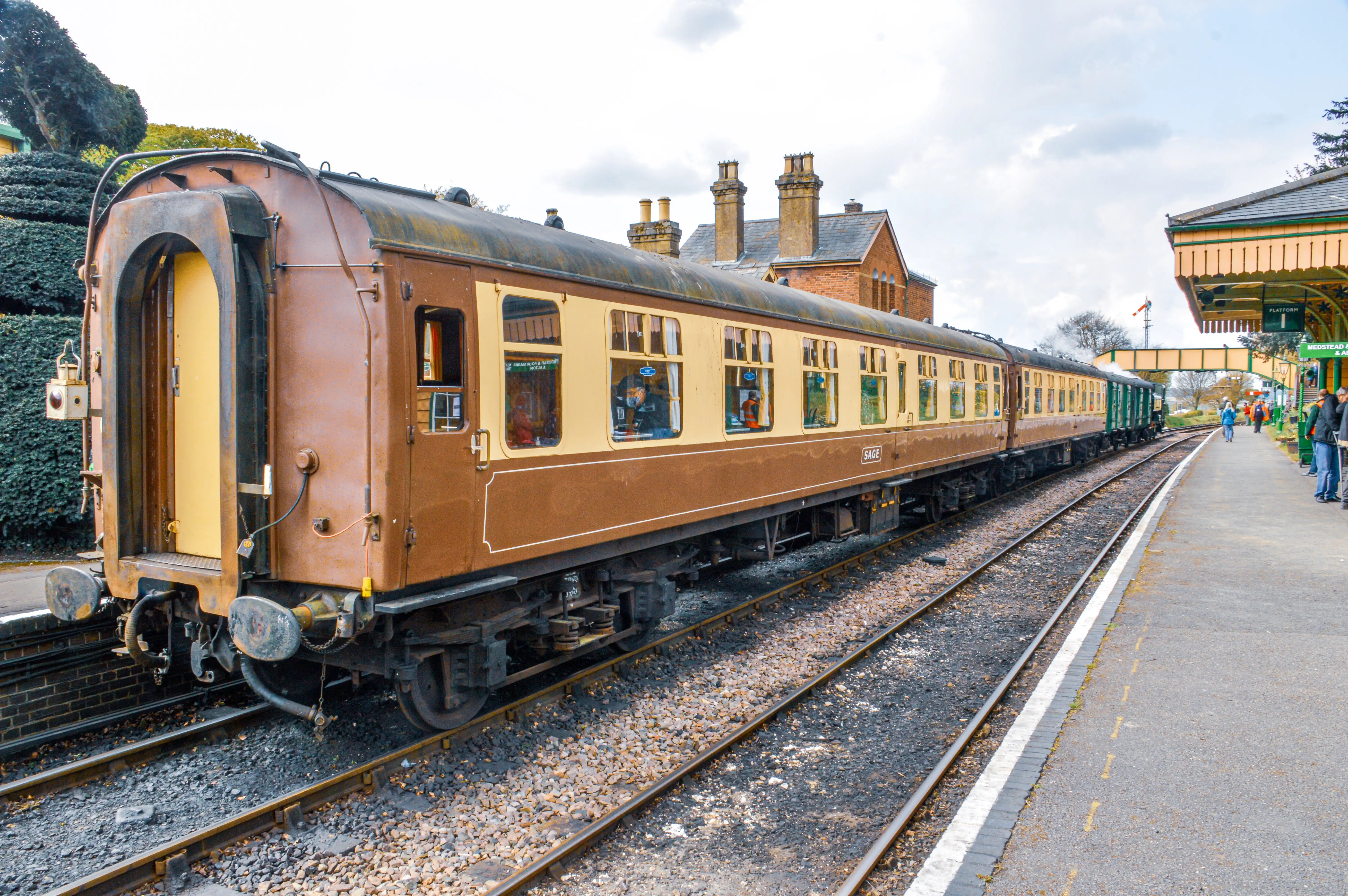 The image depicts a vintage train at a railway station. The train has a brown and cream color scheme and appears to be well-maintained. The station platform is visible on the right side, with a few people waiting. The station building has a traditional architectural style with brick chimneys and a decorative roof.
