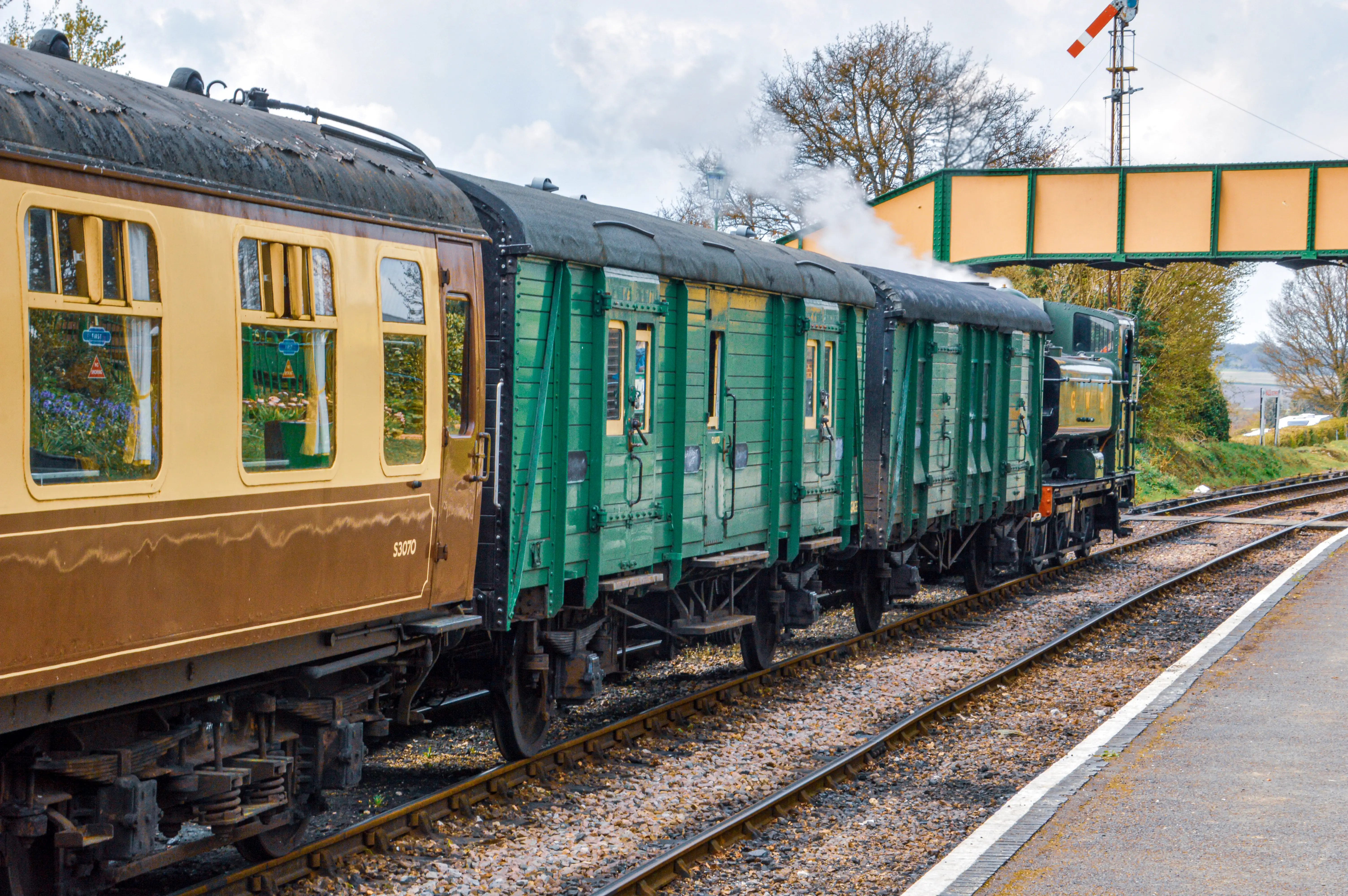 The image depicts a vintage train at a railway station. The train consists of a brown passenger car and green freight cars. Steam is visible, indicating the train is likely powered by a steam engine. The setting appears to be a rural or historical railway line, with a pedestrian bridge and some greenery in the background.