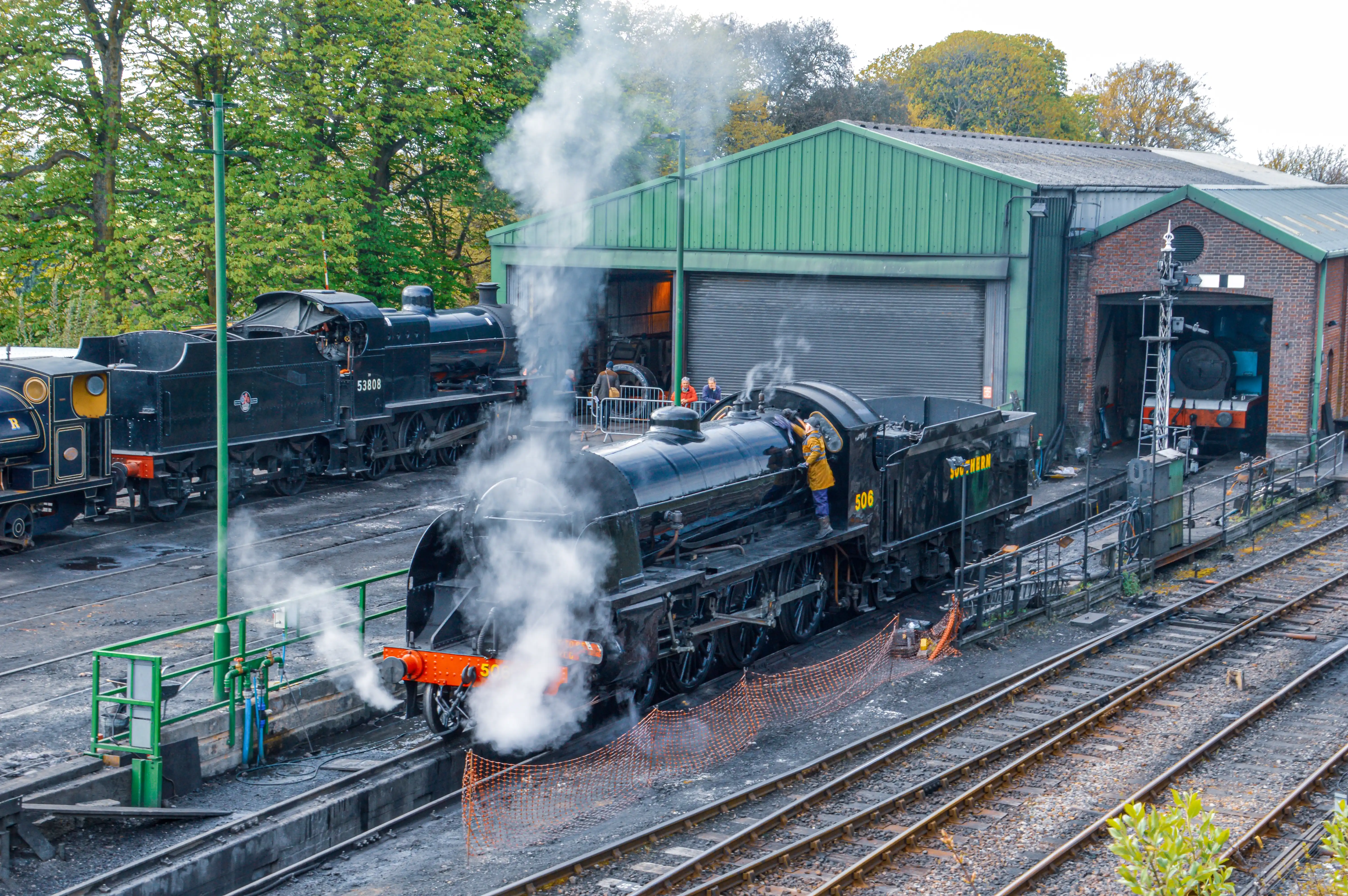 The image depicts a steam locomotive at a railway station or depot. The locomotive is emitting steam, indicating it is either preparing to depart or has just arrived. There are other locomotives and rail cars visible in the background, and the setting appears to be a maintenance or storage facility. The scene is surrounded by greenery, suggesting a semi-rural or suburban location. A person in a yellow jacket is visible on the locomotive, possibly a crew member or engineer.