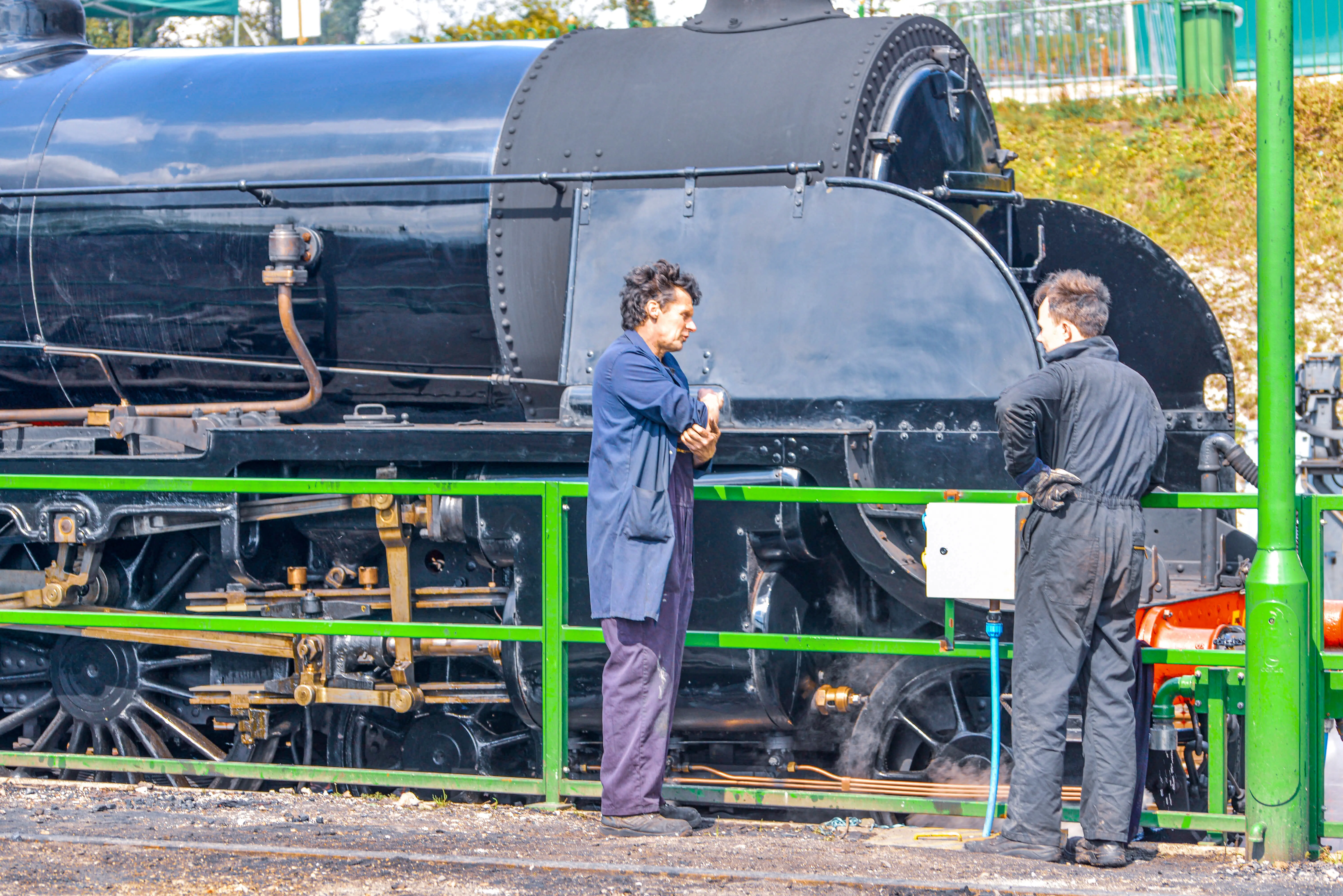 The image shows two individuals standing next to a large, black steam locomotive. One person is wearing a blue work coat and the other is in dark overalls. They appear to be engaged in conversation or inspection near the front wheels of the locomotive, which is situated on railway tracks. The scene is set outdoors, with a green railing and some greenery visible in the background.