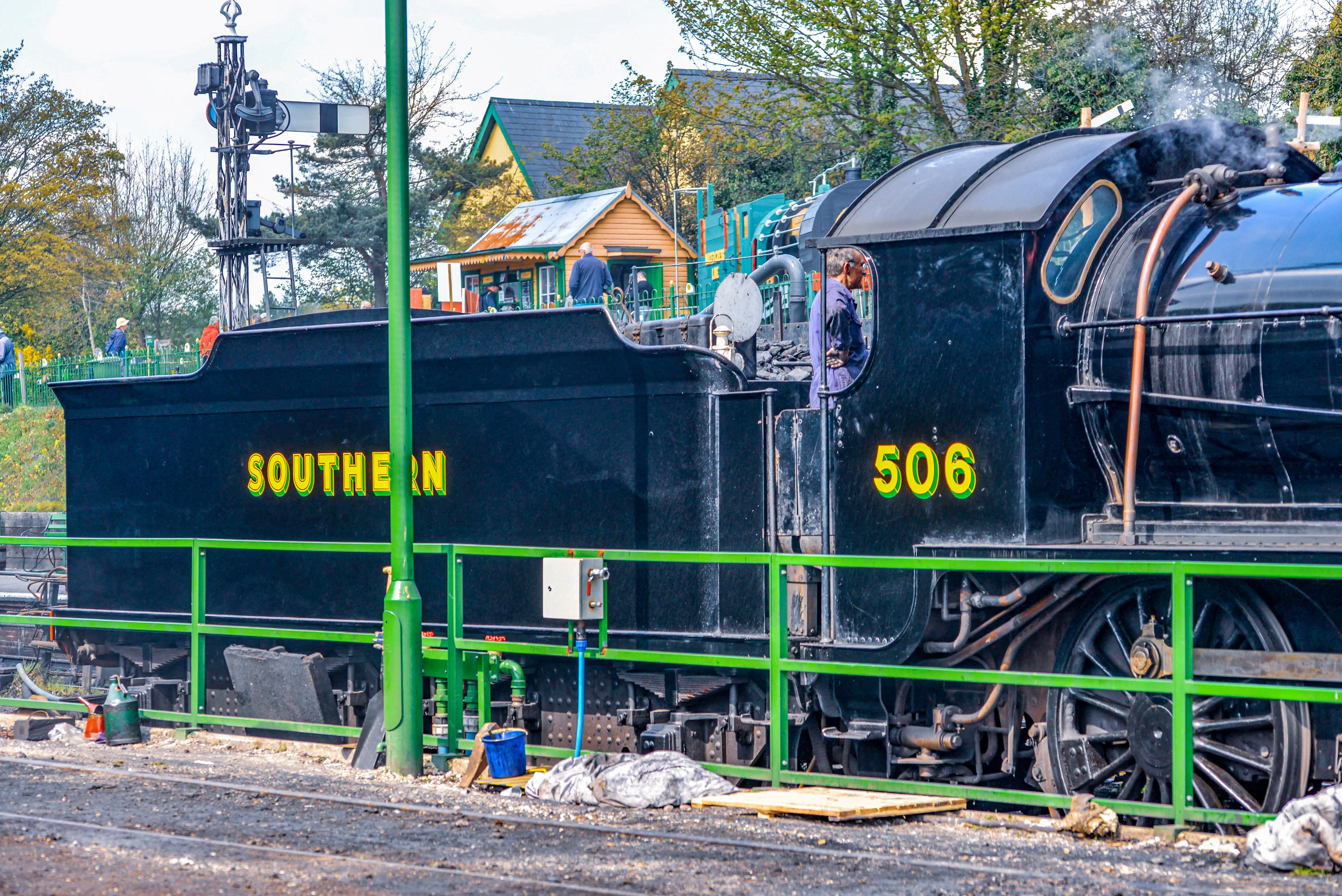 The image depicts a black steam locomotive with the word 'SOUTHERN' and the number '506' prominently displayed on its side. The locomotive is stationed on railway tracks, with steam visibly emanating from its engine. Surrounding the train are various railway infrastructure elements, including signals and fencing. In the background, there are small buildings and greenery, indicating a railway station or a train yard setting.