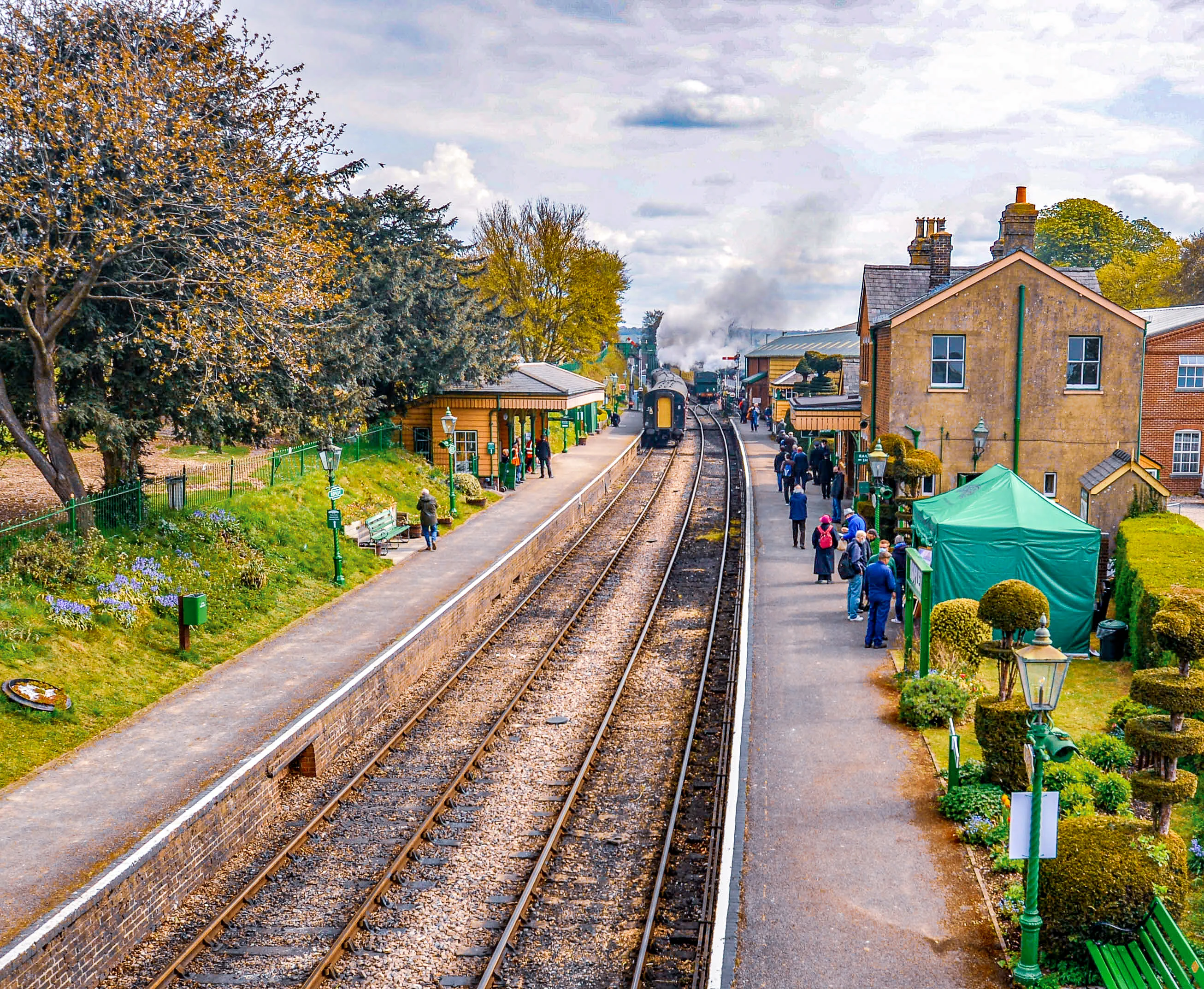 The image depicts a quaint train station with a vintage steam train arriving. The station is surrounded by lush greenery and well-maintained gardens. People are seen walking along the platform, and the architecture of the station buildings has a classic, historical look. The overall scene suggests a peaceful, nostalgic atmosphere, possibly in a rural or heritage railway setting.