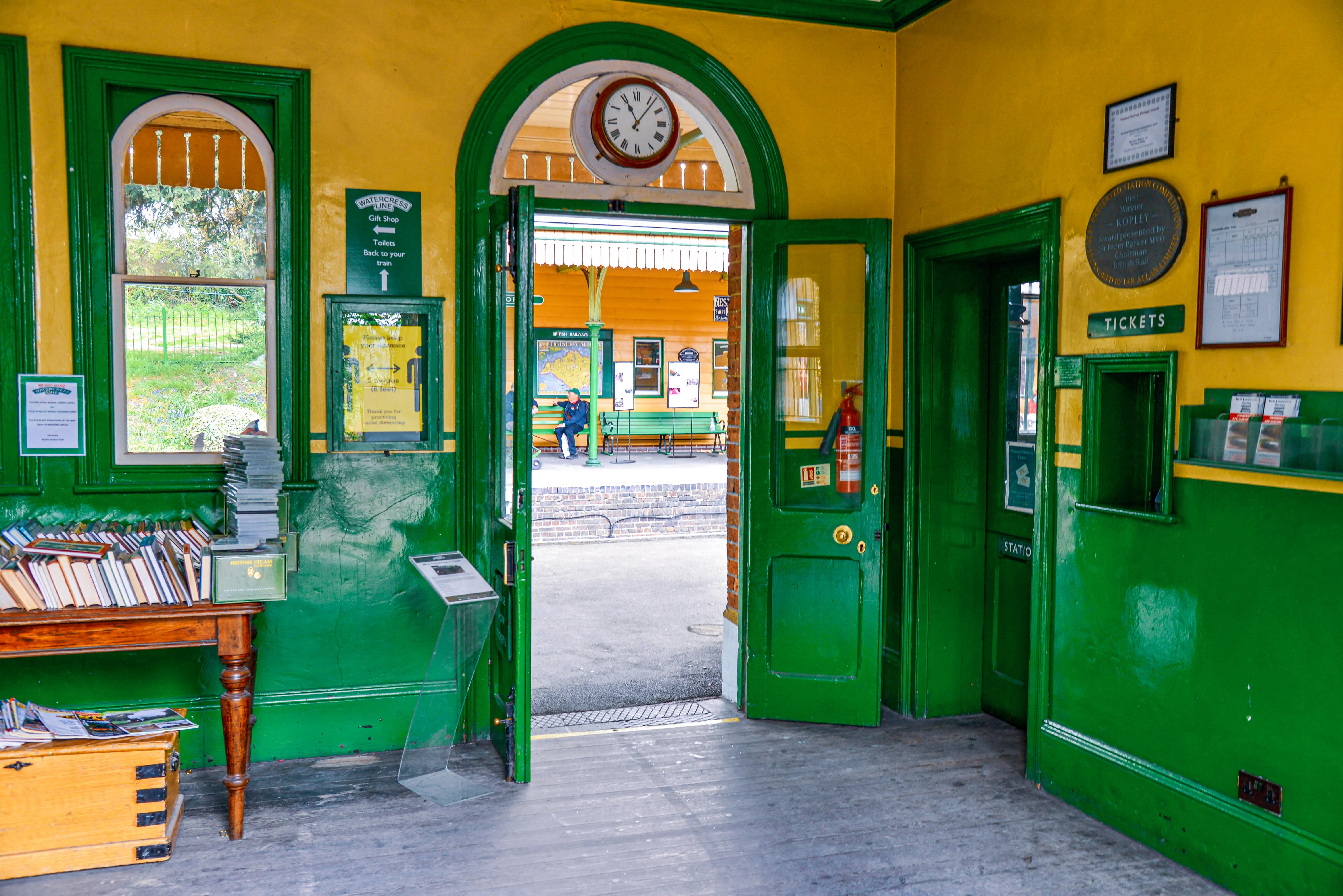 The image depicts the interior of a quaint, vintage train station. The station features green and yellow decor with a clock above the open double doors leading to the platform. Inside, there is a small wooden table with books and pamphlets, and various signs and notices on the walls. The overall atmosphere is nostalgic and well-preserved, reflecting an older architectural style.
