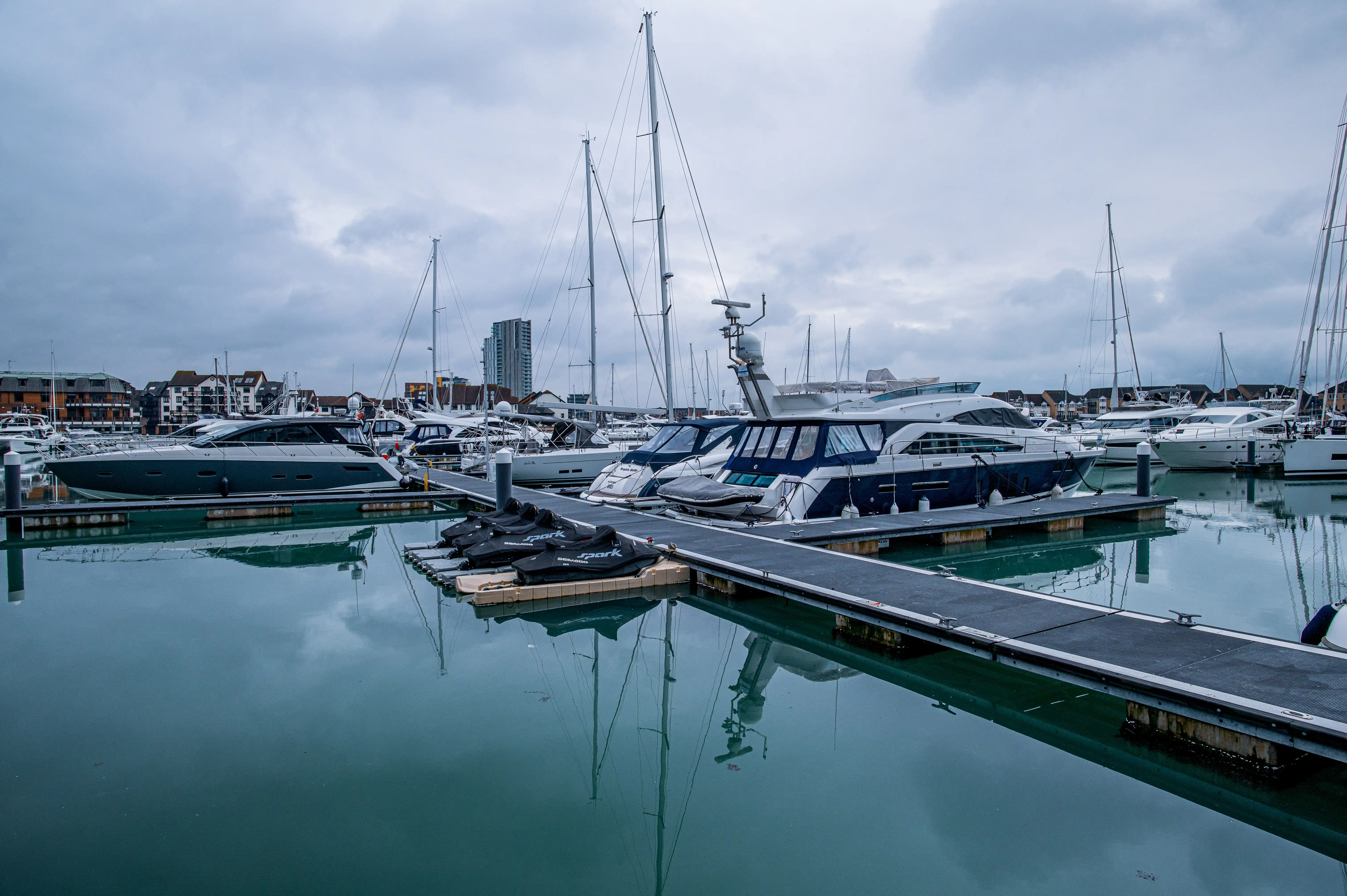 This image captures a peaceful marina filled with various boats, from sleek motor yachts to tall-masted sailing vessels. The cloudy sky casts a serene, muted light over the scene, with the reflections of the boats and the sky shimmering on the calm water. Several jet skis are neatly docked along a wooden walkway, adding a touch of recreational activity to the otherwise tranquil setting. In the background, buildings line the shore, suggesting a bustling town or city nearby.