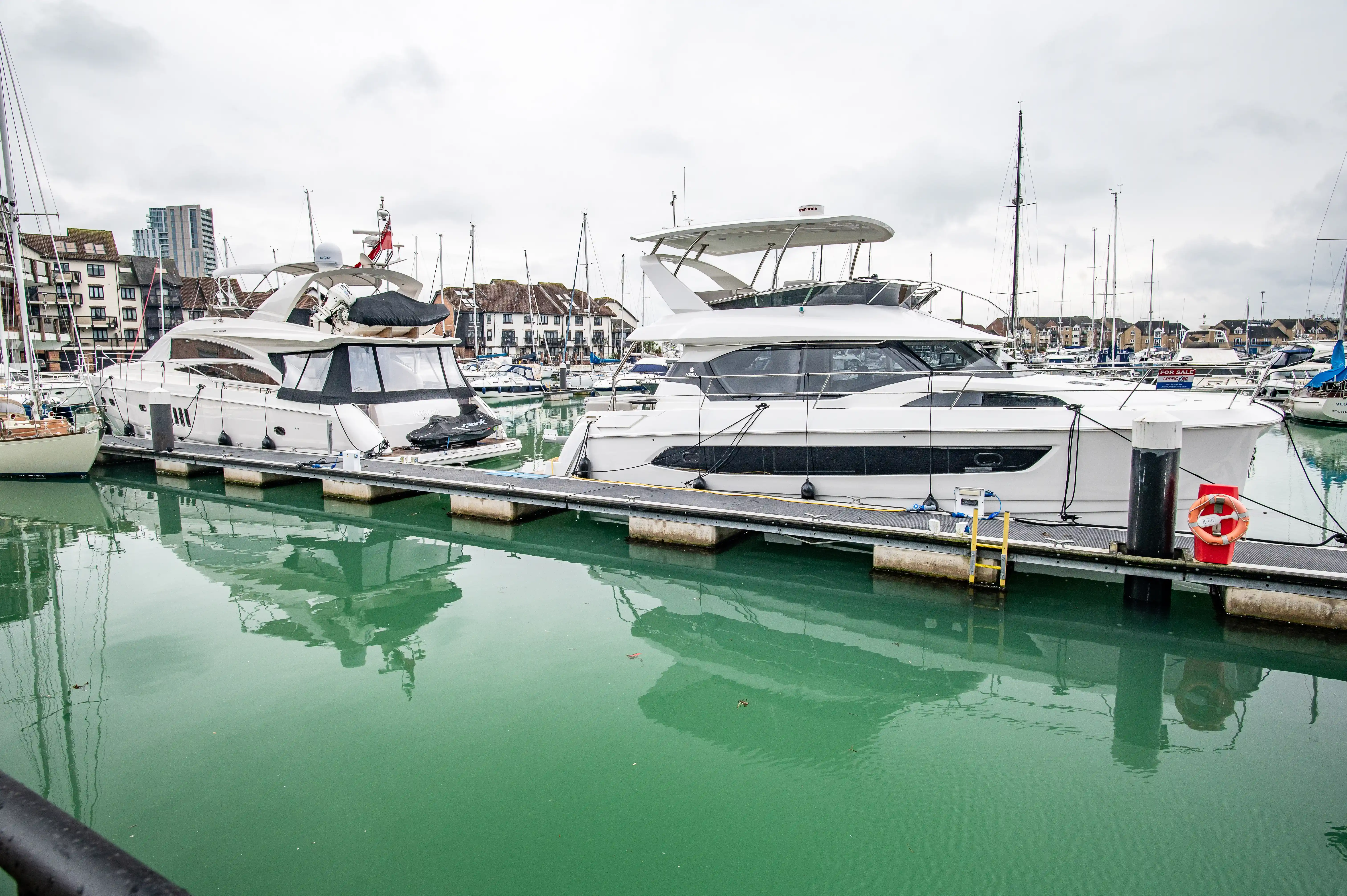 The image depicts a marina with several luxury yachts docked at the pier. The yachts are modern and well-maintained, featuring large windows and sleek designs. The water in the marina is calm and reflects the boats, creating a serene atmosphere. In the background, there are buildings and other boats, indicating a bustling yet orderly harbor. The overall scene suggests a high-end, well-kept marina.