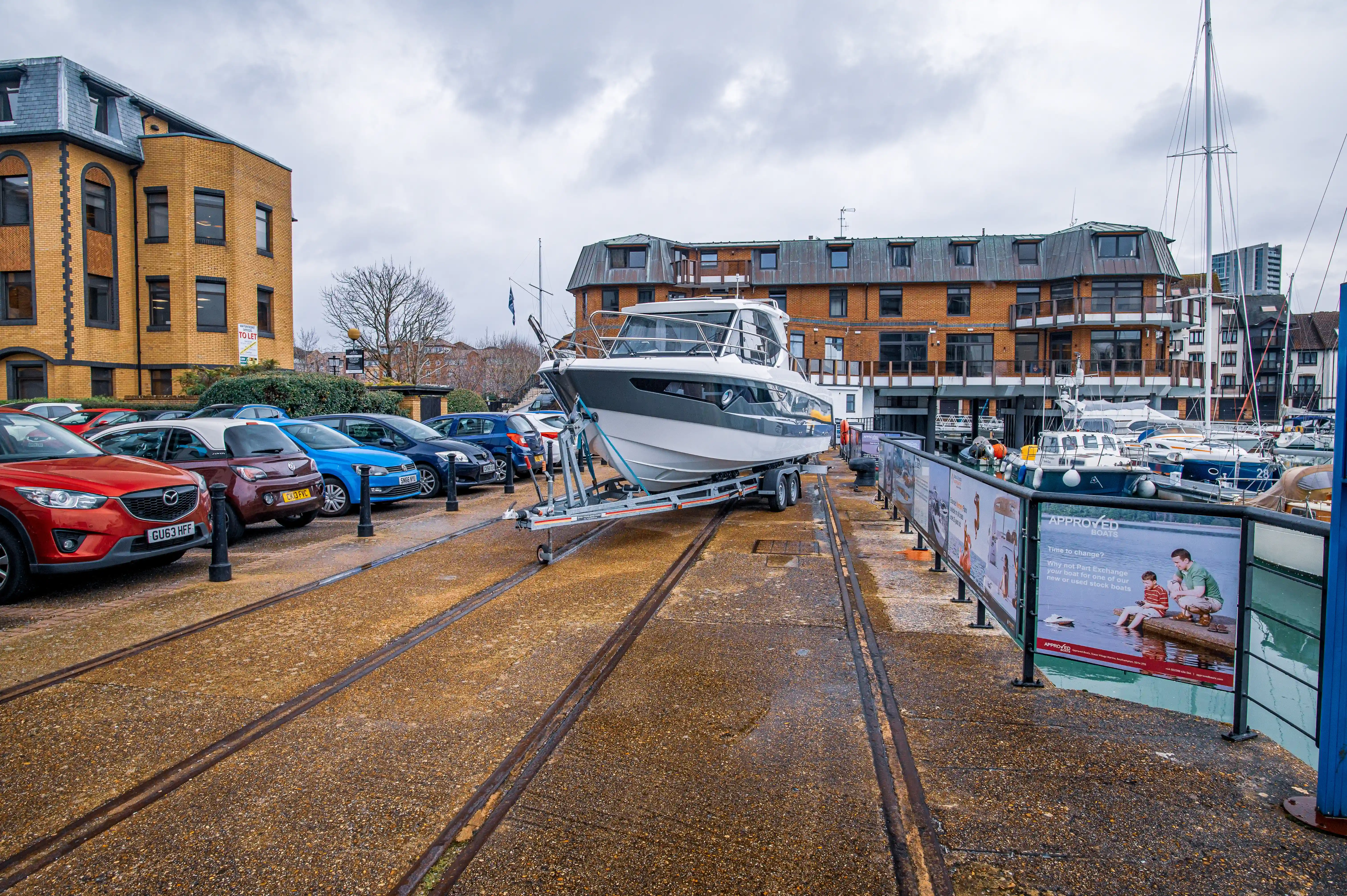 The image depicts a marina area with a boat being transported on a trailer, surrounded by parked cars and buildings. The scene includes railway tracks in the foreground and advertisements for boats in the background. The weather appears overcast, and the ground is wet, suggesting recent rain.