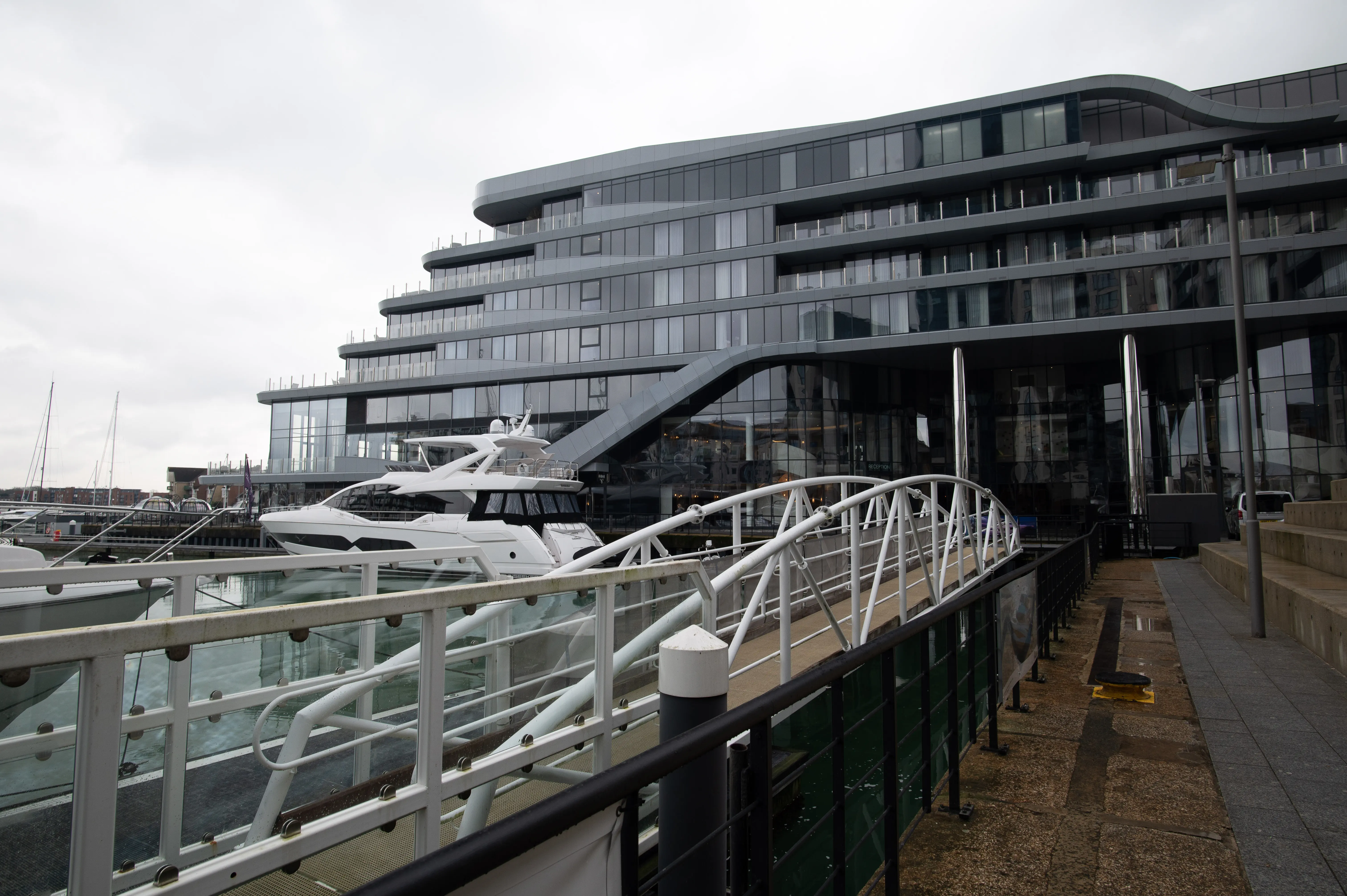 The image depicts a modern marina with a multi-story building featuring a unique, wavy architectural design. The building has large glass windows and is situated directly on the waterfront. Several yachts and boats are docked in the marina, with a prominent white yacht in the foreground. The area is enclosed by railings, and the overall scene suggests a luxurious and contemporary setting.
