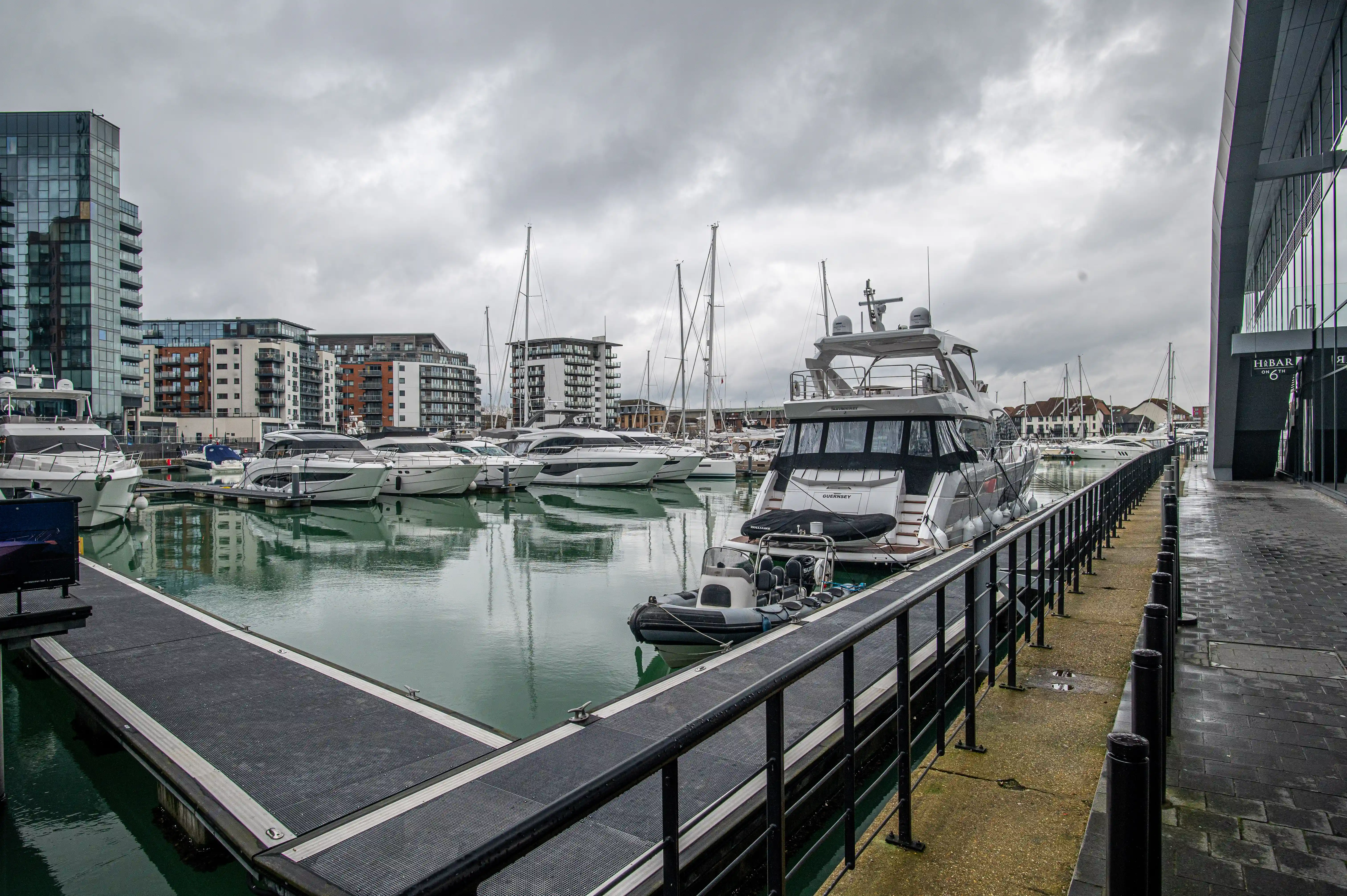 The image depicts a marina with several docked boats and yachts. The marina is surrounded by modern buildings and apartments. The weather appears to be overcast, and the water in the marina is calm. There is a walkway along the edge of the marina with railings, and a small inflatable boat is tied to one of the larger yachts.