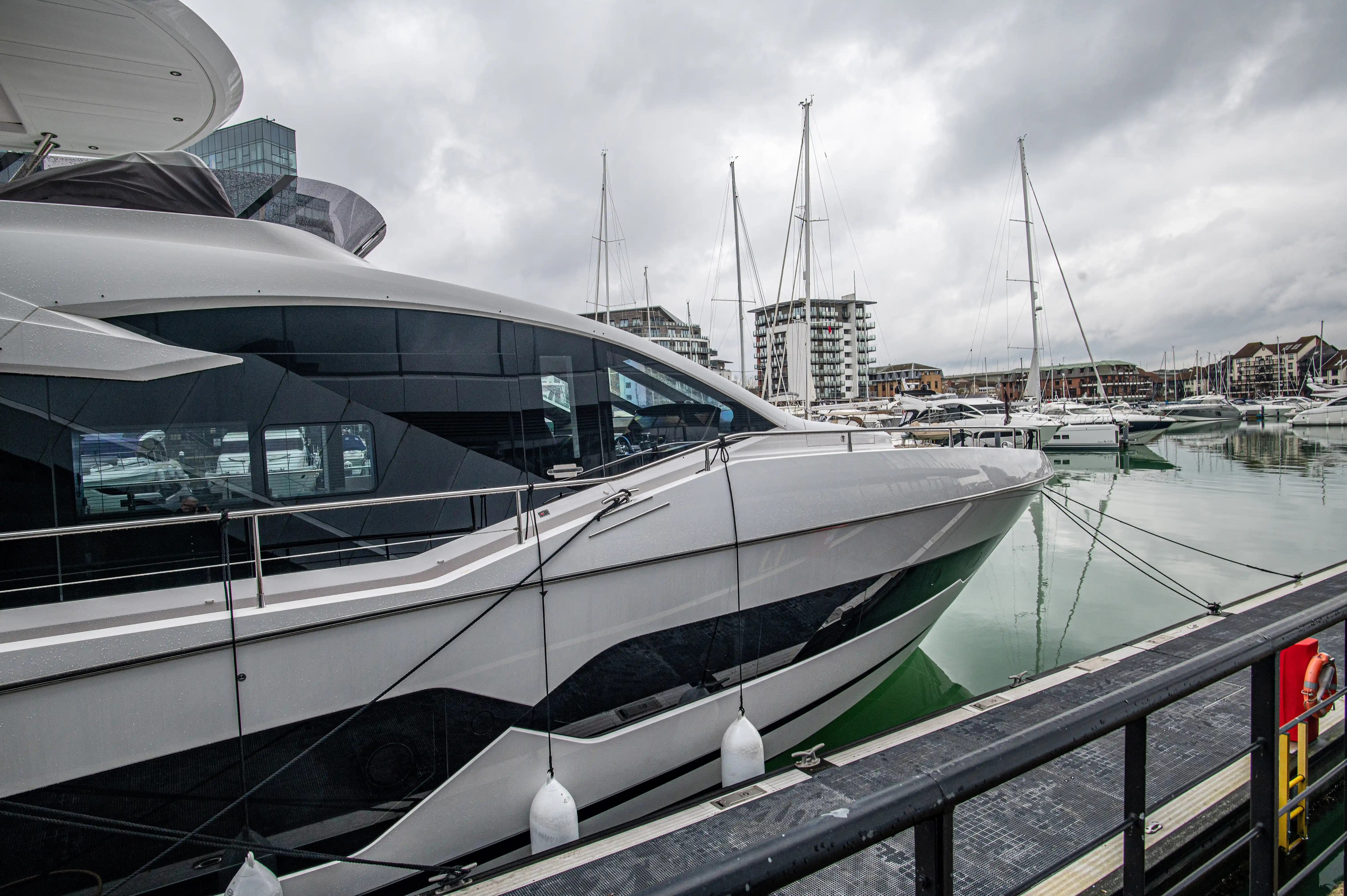 The image depicts a modern marina with several luxury yachts docked. The yachts have sleek, contemporary designs with large windows and streamlined exteriors. The marina is surrounded by modern buildings, and the scene is set on a cloudy day, with the water in the marina being calm and reflecting the boats and surroundings.