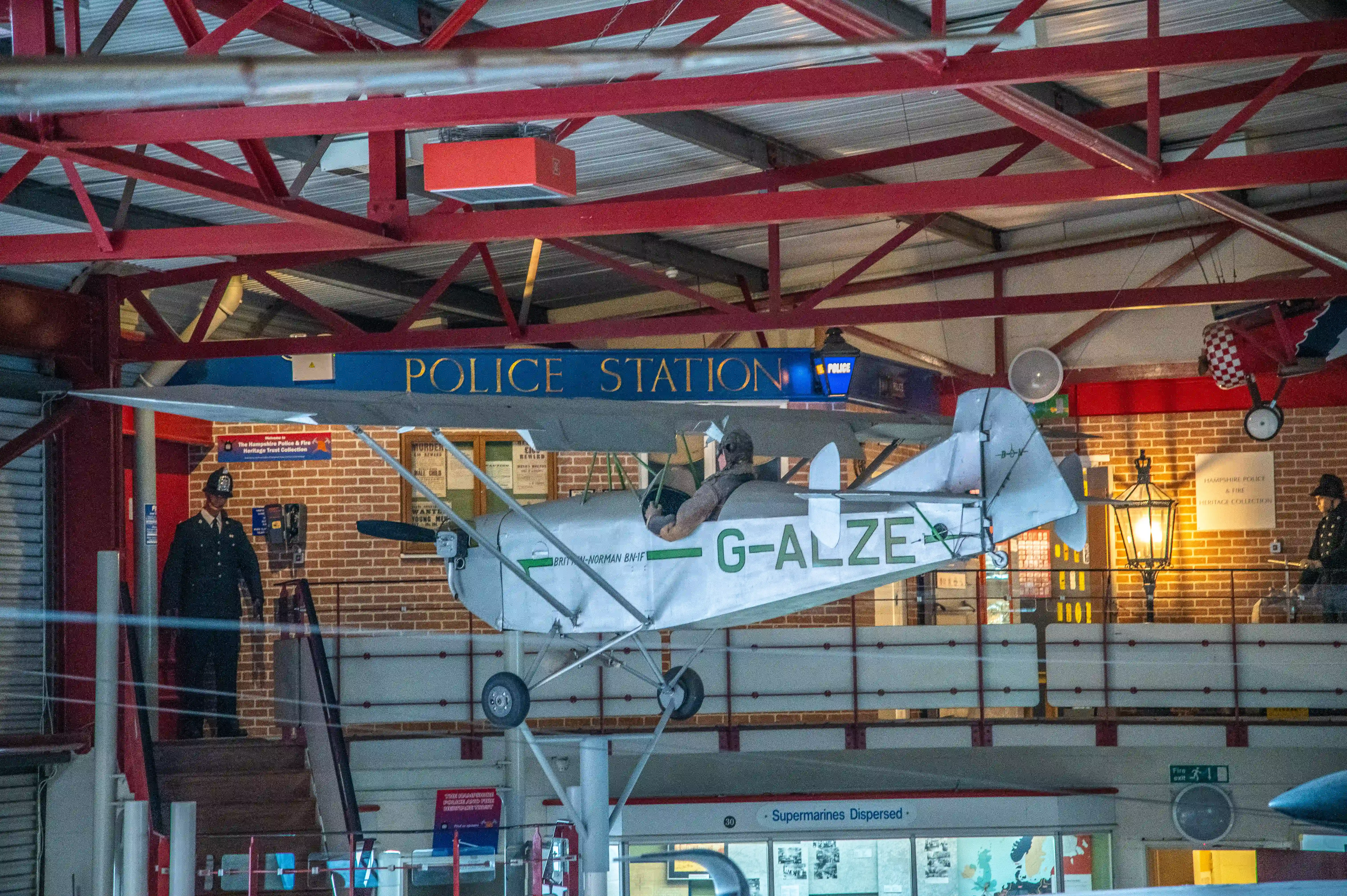 The image depicts an indoor exhibit at a police station. A vintage biplane labeled 'G-ALZE' is suspended from the ceiling, and the station features a sign that reads 'POLICE STATION.' There are two police officers in uniform, and various displays and signs related to police and fire heritage can be seen in the background.