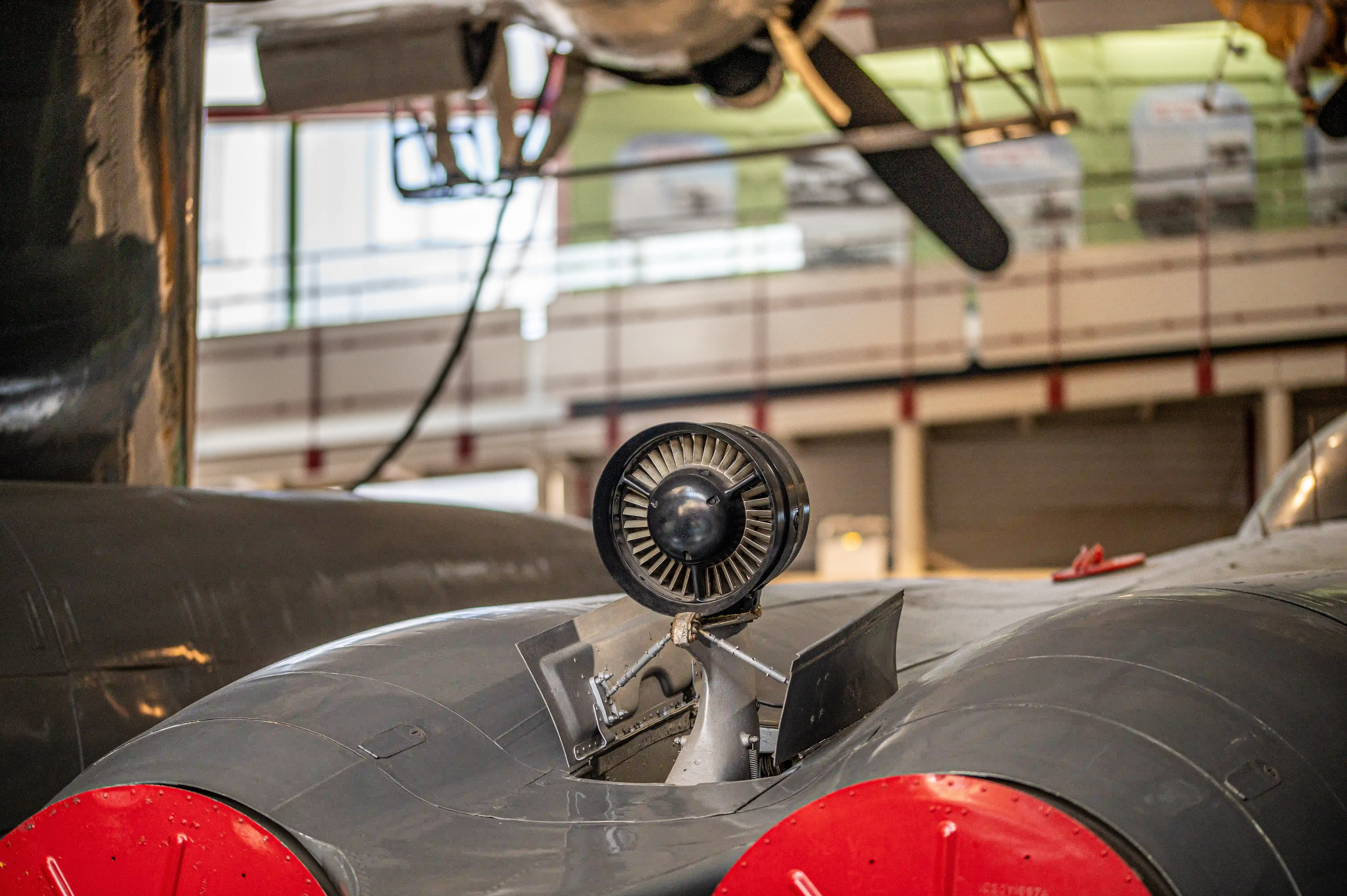 The image depicts the interior of an aircraft, focusing on the engine and its components. The view is from inside the cockpit, looking out through the propeller. The propeller has a central hub with three blades, and a round, metallic object, likely a cowl flap, is prominently visible on the engine. The background shows a blurred view of a building, suggesting the aircraft is in a hangar or similar setting.