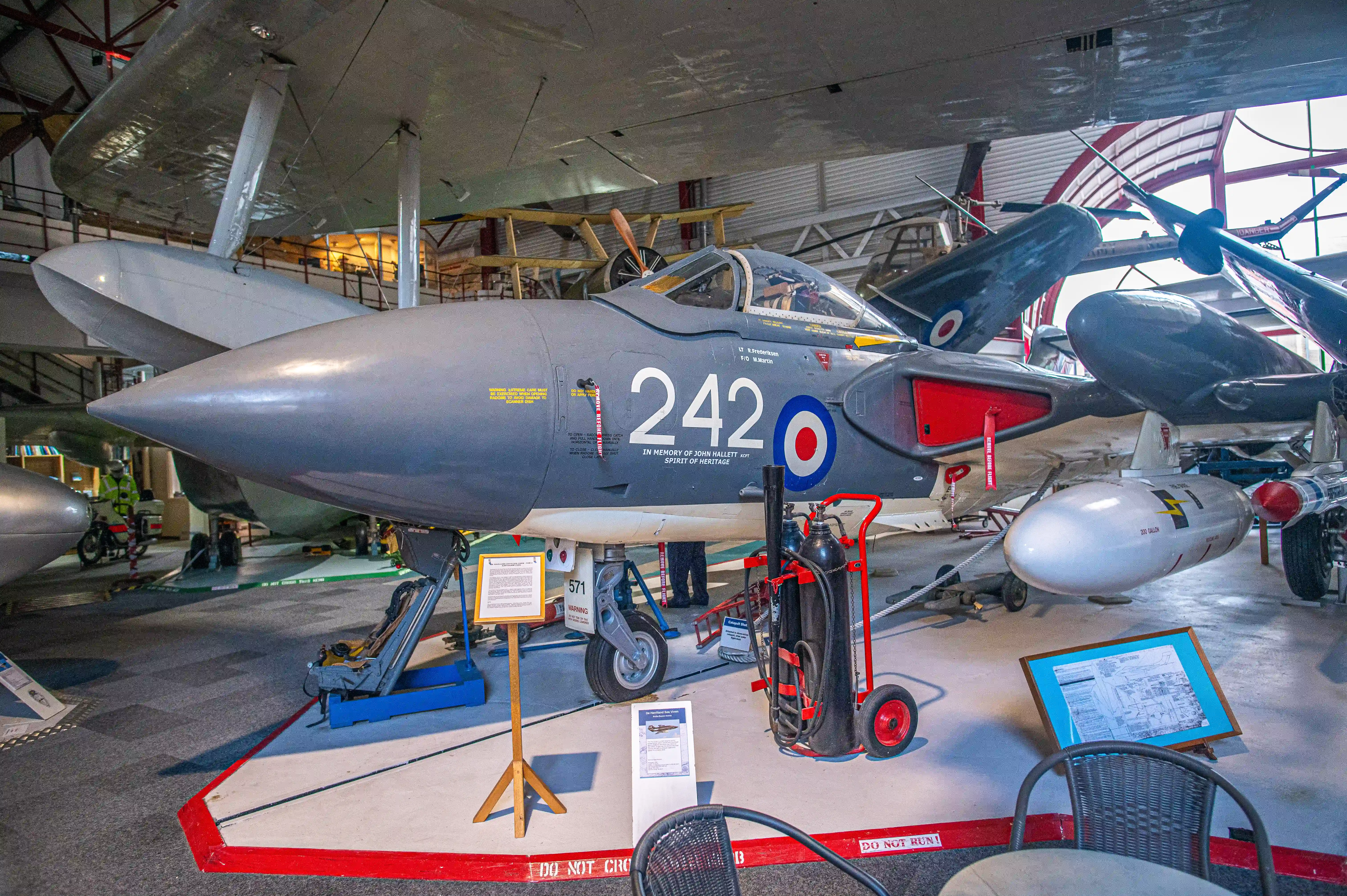 The image depicts a vintage military aircraft, specifically a jet fighter, on display in a museum. The aircraft has the number 242 painted on its fuselage along with a roundel insignia. Various equipment and informational placards are placed around the aircraft, providing details about its history and specifications. The setting appears to be an indoor aviation museum with other aircraft and exhibits visible in the background.