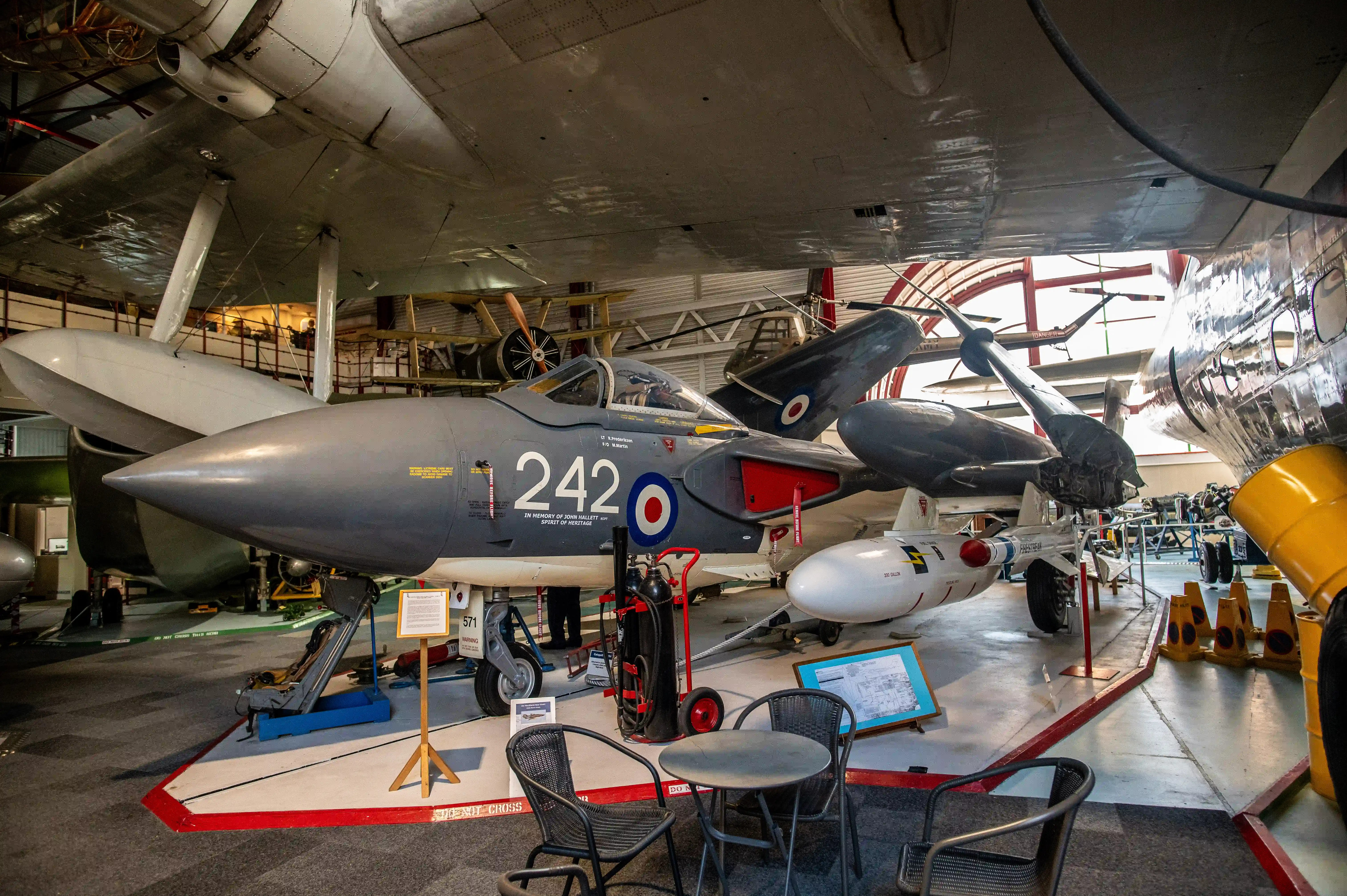 The image depicts an aircraft museum exhibit featuring a grey fighter jet marked with the number 242 and a roundel insignia. The jet is displayed indoors alongside other aircraft and aviation equipment. There are informational placards and a small seating area with a table in front of the jet. The setting appears to be well-lit with natural light streaming in from large windows.