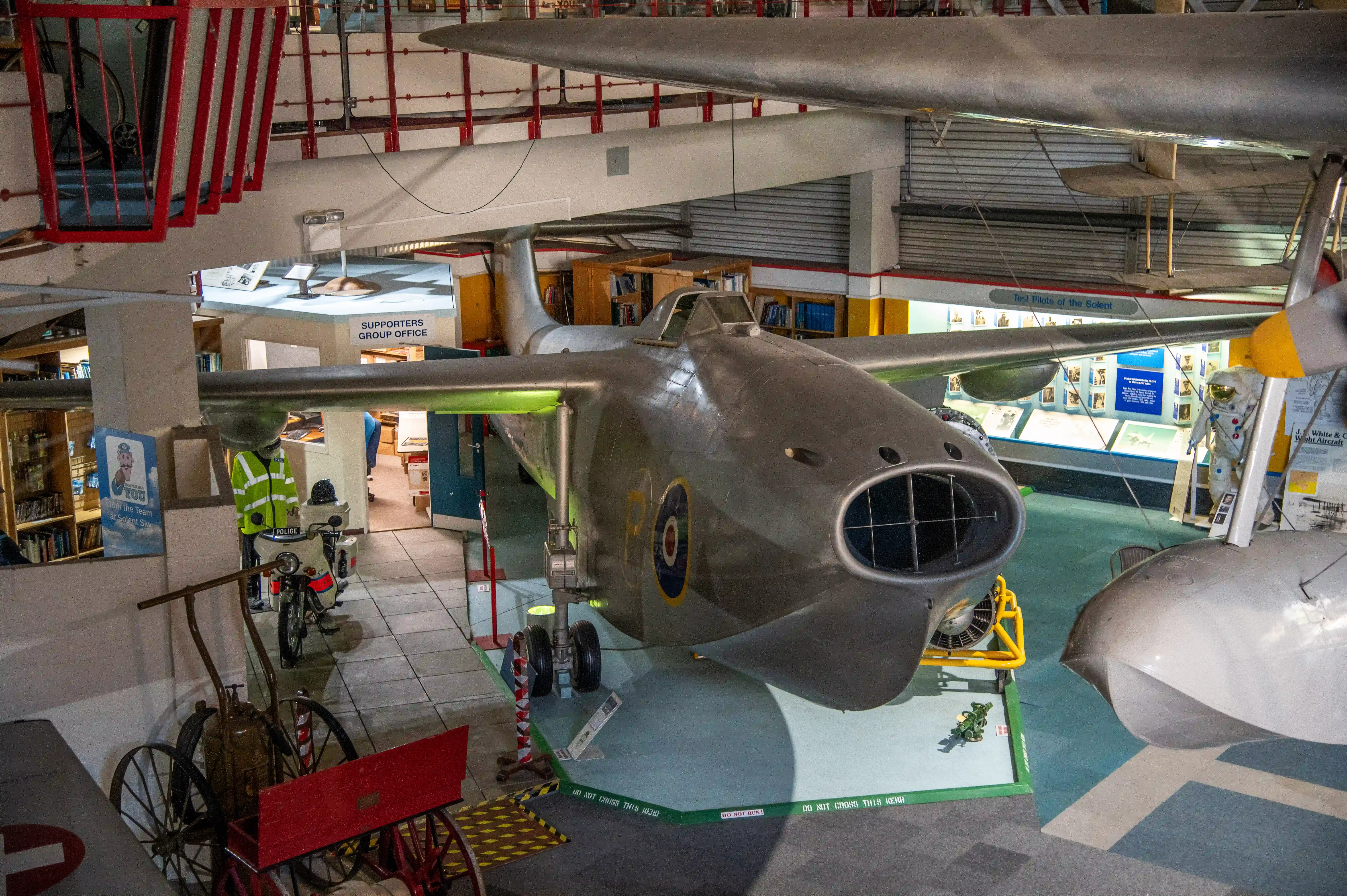 The image depicts an indoor museum exhibit featuring a large, vintage military aircraft. The aircraft is prominently displayed in the center of the room, with its nose and cockpit clearly visible. Surrounding the aircraft are various other exhibits and informational displays, including a motorcycle, a bicycle, and several posters and signs. The room is well-lit and has a high ceiling with exposed beams and red accents. There is a person in a high-visibility jacket standing near the aircraft, possibly a museum staff member or security personnel.