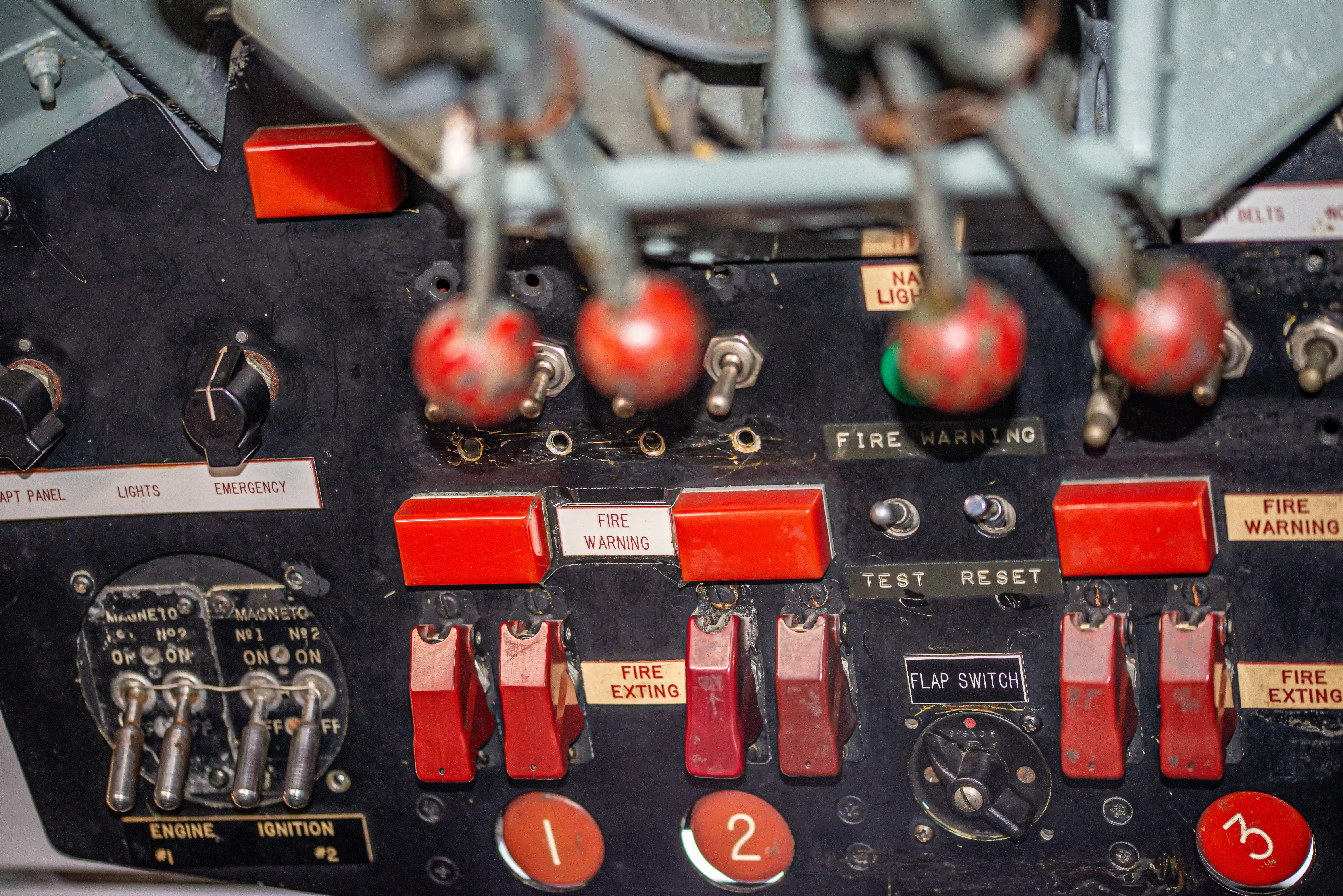 The image shows a close-up of an aircraft's control panel, featuring various switches and indicators. Key elements include fire warning switches, fire extinguishing controls, engine ignition switches, and a flap switch. The panel also has labels for different functions such as lights, emergency, and magneto controls. The switches are predominantly red, indicating their importance in emergency situations. The overall layout is organized with clear labeling to assist the pilot in managing critical systems during flight.