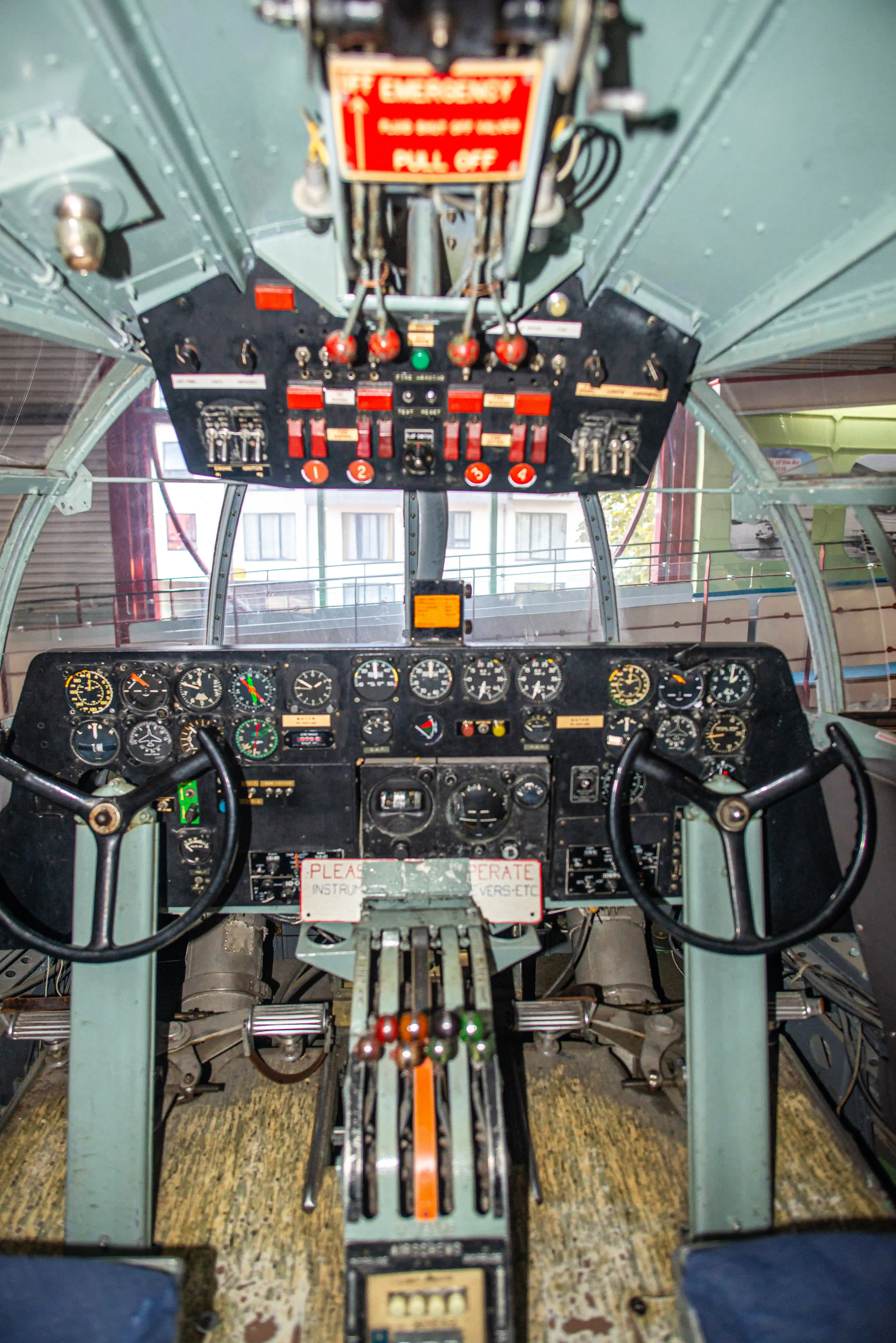The image shows the interior cockpit of an aircraft, featuring a detailed view of the control panels and instruments. The cockpit is equipped with various gauges, switches, and control sticks, indicating it is likely from an older aircraft model. There are signs instructing pilots and visitors, such as 'Emergency - Push to Open' and 'Please Instruct Before Manipulating Controls - Versatile.' The overall setup suggests a historical or training environment, possibly in a museum or an aviation exhibit.