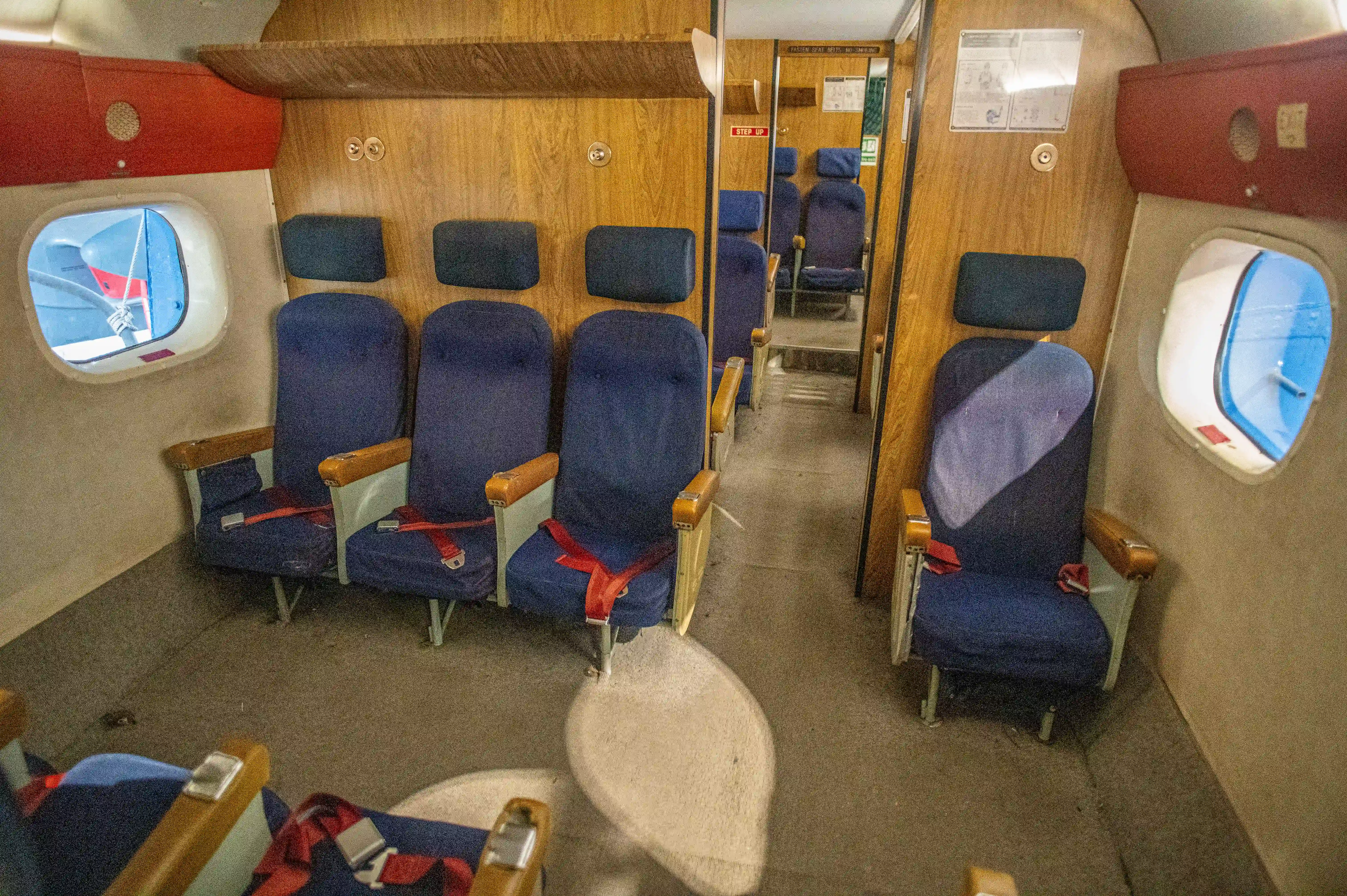 The image shows the interior of an airplane with rows of blue seats that have wooden armrests and headrests. The seats are arranged in a 2-2 configuration, with two seats on either side of the aisle. Each seat is equipped with a red seatbelt. The walls are wooden, and there are round windows on either side of the plane. The floor is carpeted, and there are various items, including a blanket and a bag, on the seats and the floor.