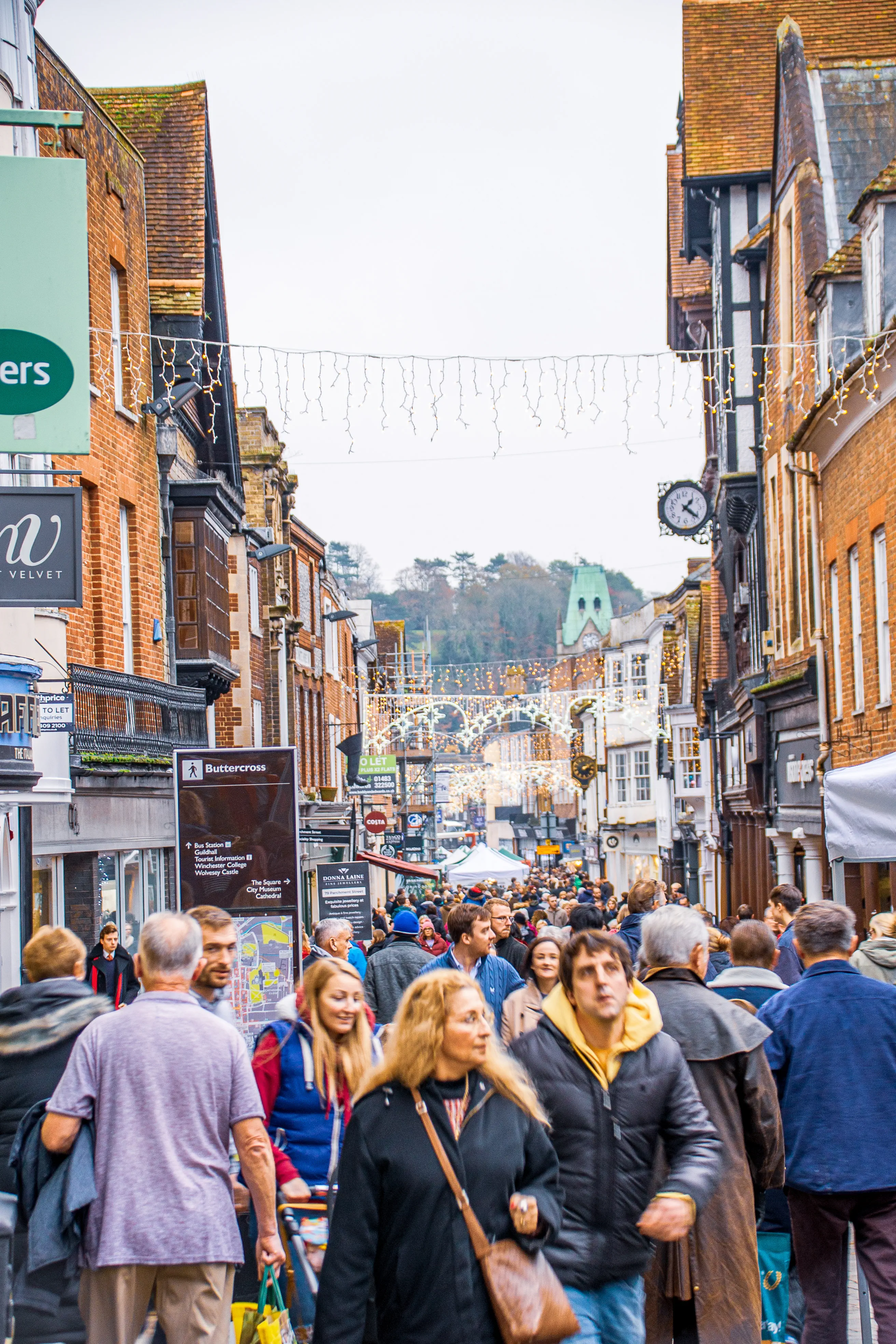 The image depicts a bustling street scene in what appears to be a historic town. The street is crowded with people, suggesting a busy day, possibly during a holiday season due to the festive lights strung above. Shops and buildings line both sides of the street, and a sign for Buttercross is visible, indicating a notable landmark. The architecture of the buildings is traditional, adding to the charm of the scene. The overall atmosphere is lively and vibrant.