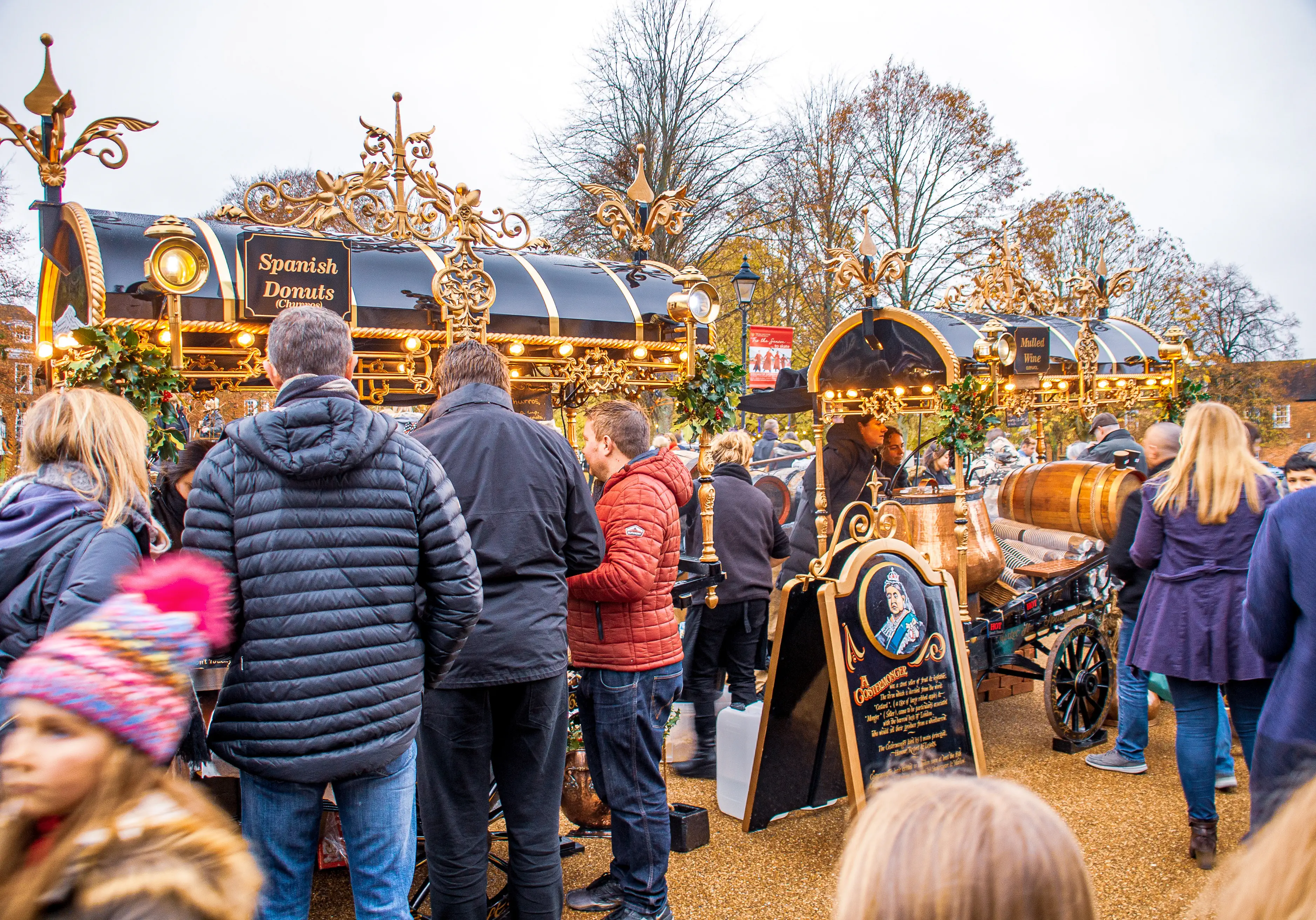 The image depicts a bustling outdoor market scene with people dressed in warm clothing. There are ornate stalls offering food and beverages, including Spanish donuts and mulled wine. The stalls are decorated with festive lights and garlands, and there is a sign with a historical figure. The setting appears to be a festive or holiday market, likely during a cooler season.