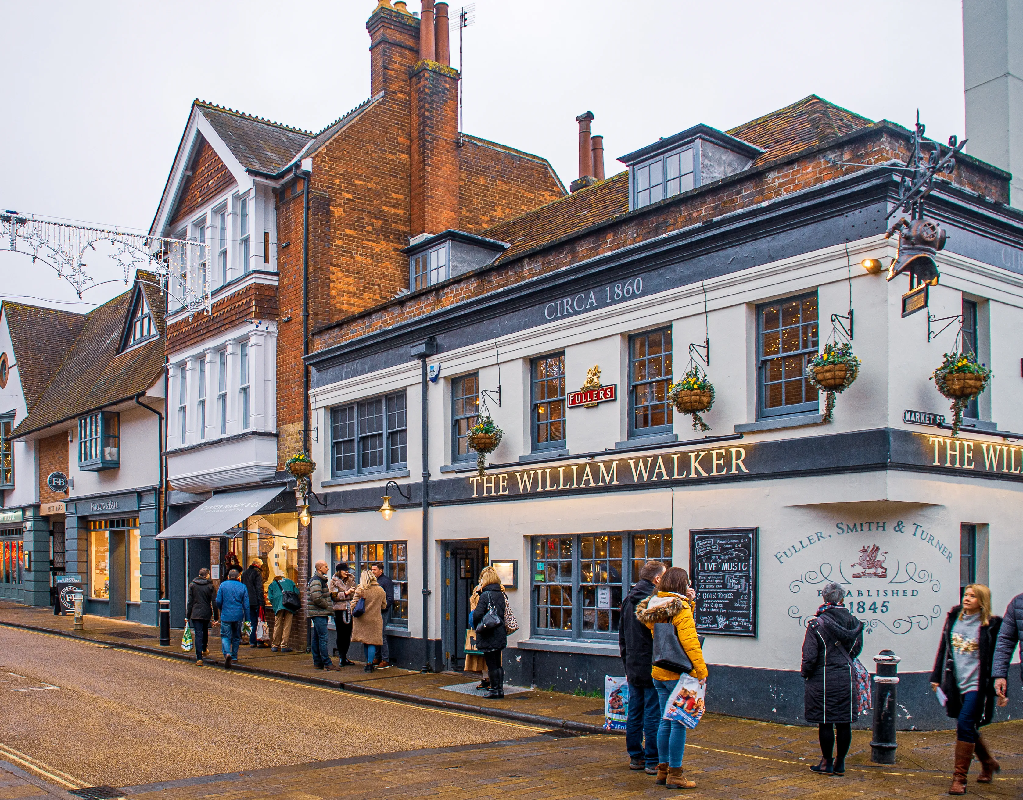 The image depicts a lively street scene centered around a historic pub named 'The William Walker,' established circa 1860. The building features traditional architecture with hanging flower baskets and a sign indicating it is associated with Fuller, Smith & Turner, established in 1845. People are seen walking, socializing, and entering the pub, contributing to a bustling and inviting atmosphere. The street is lined with other shops and buildings, adding to the quaint and charming ambiance of the area.