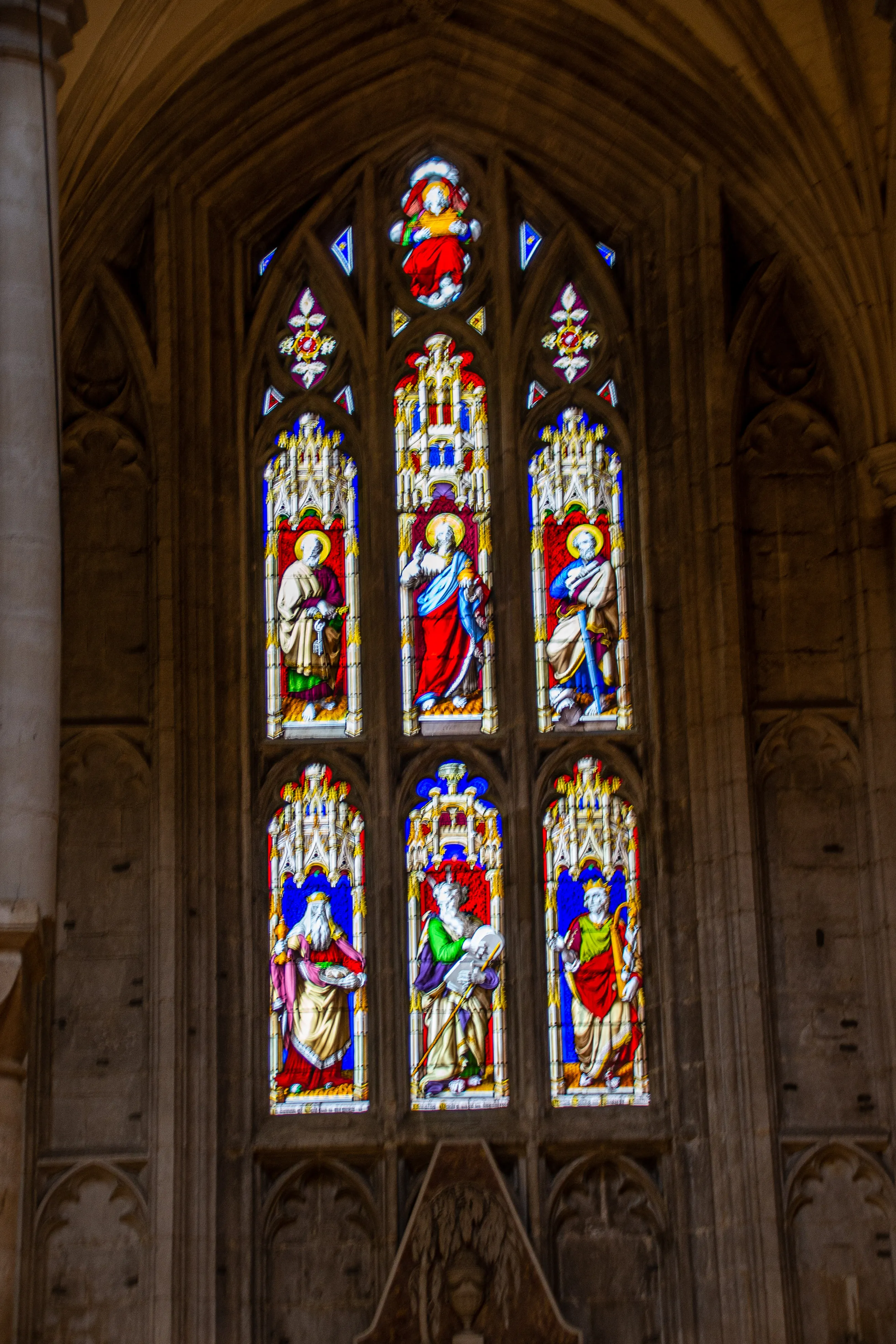 The image depicts a stained glass window in a Gothic architectural style, commonly found in churches or cathedrals. The window is divided into six panels, each featuring a religious figure with halos, adorned in traditional robes. The figures are set against vibrant, colorful backgrounds with intricate designs and patterns. The craftsmanship highlights detailed facial expressions and postures, indicative of historical and religious significance.
