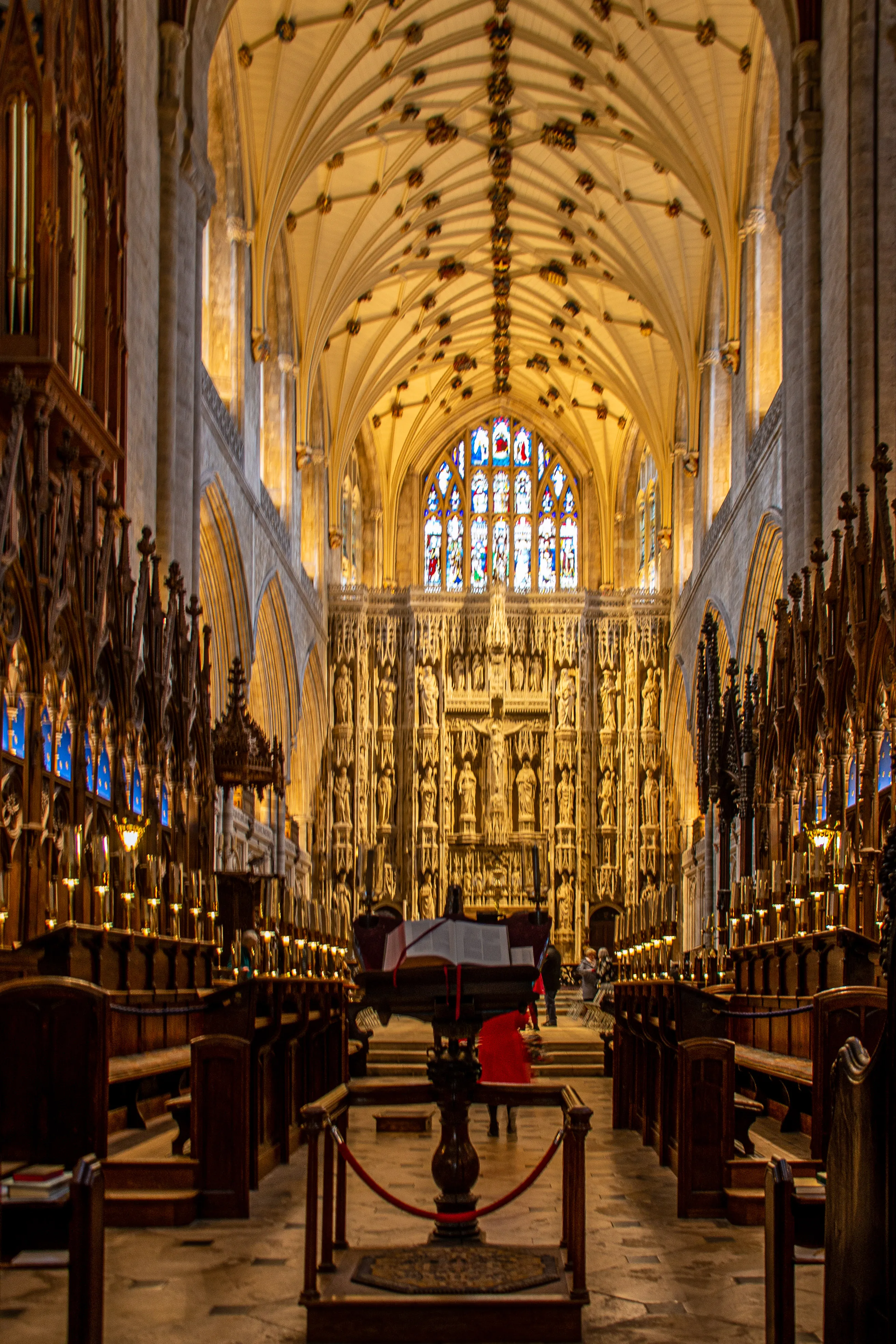 The image depicts the interior of a grand cathedral, showcasing intricate architectural details. The view is directed towards the altar, which is adorned with elaborate carvings and golden decorations. Stained glass windows line the upper walls, allowing colored light to filter into the space. Wooden pews are arranged in rows, leading up to the altar, with a central pulpit prominently positioned. The overall ambiance is one of reverence and historical significance.
