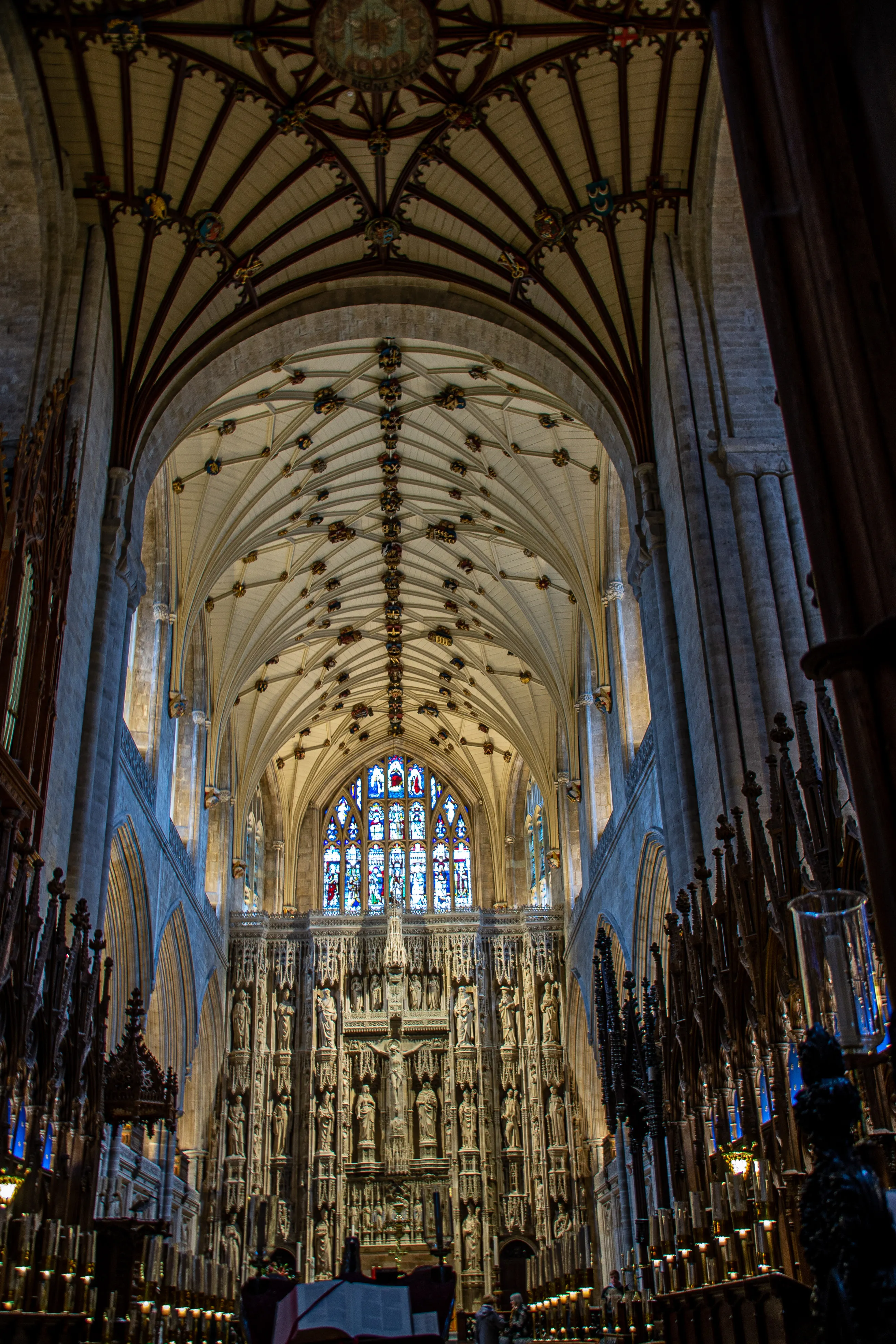 The image depicts the interior of a grand cathedral, showcasing intricate Gothic architecture. The ceiling features ribbed vaults with ornate bosses, and the space is illuminated by natural light filtering through stained glass windows. The focal point is an elaborate stone screen adorned with numerous statues, likely depicting various religious figures. The overall atmosphere is one of reverence and historical significance, emphasizing the detailed craftsmanship and architectural beauty of the cathedral.