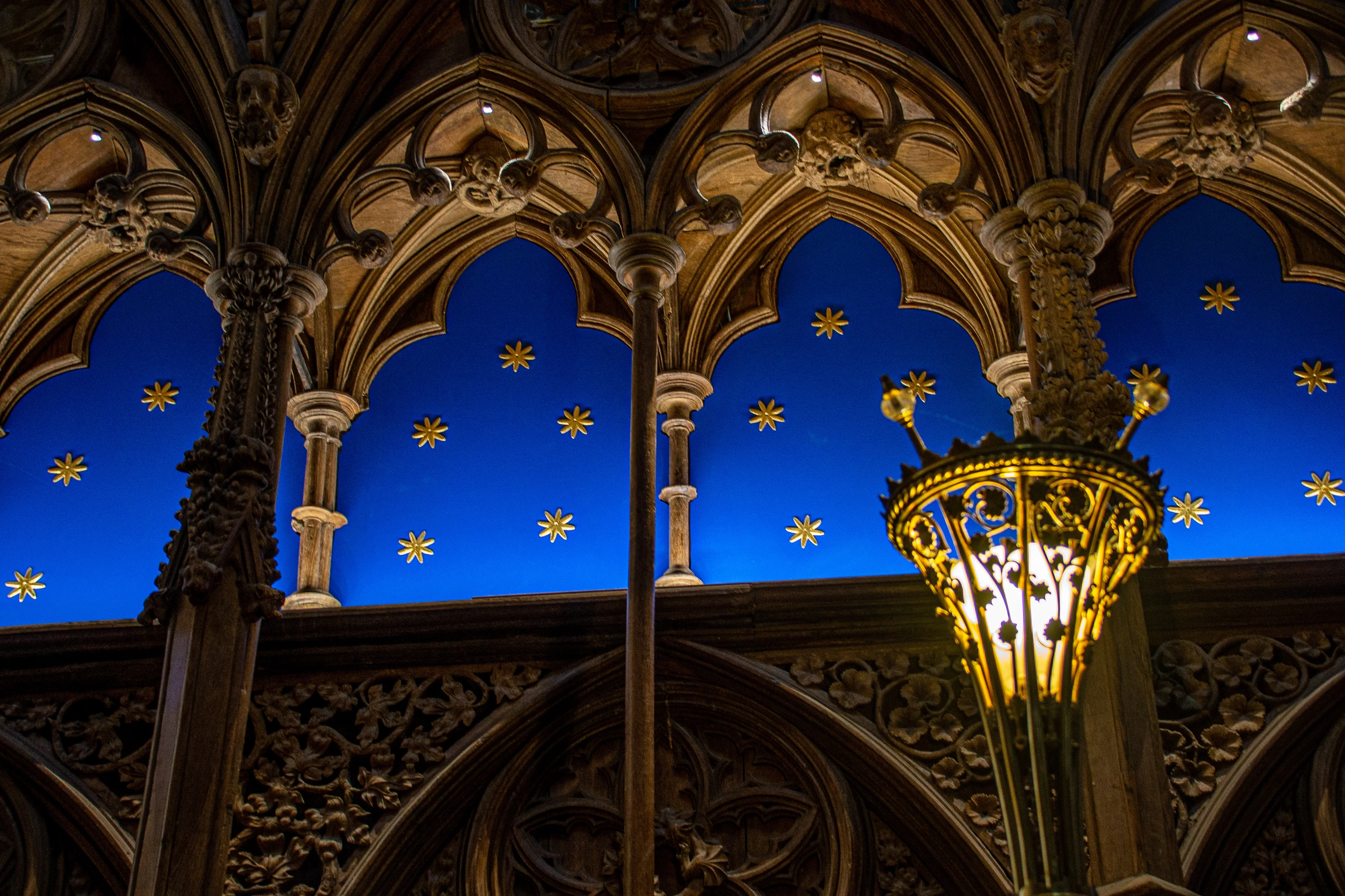 The image depicts an intricately designed interior space, likely part of a historic or religious building. It features ornate arches and columns adorned with detailed carvings and sculptures. The ceiling is painted a deep blue with golden stars, creating a night sky effect. A prominent, illuminated chandelier adds to the grandeur of the scene, highlighting the architectural beauty and craftsmanship.