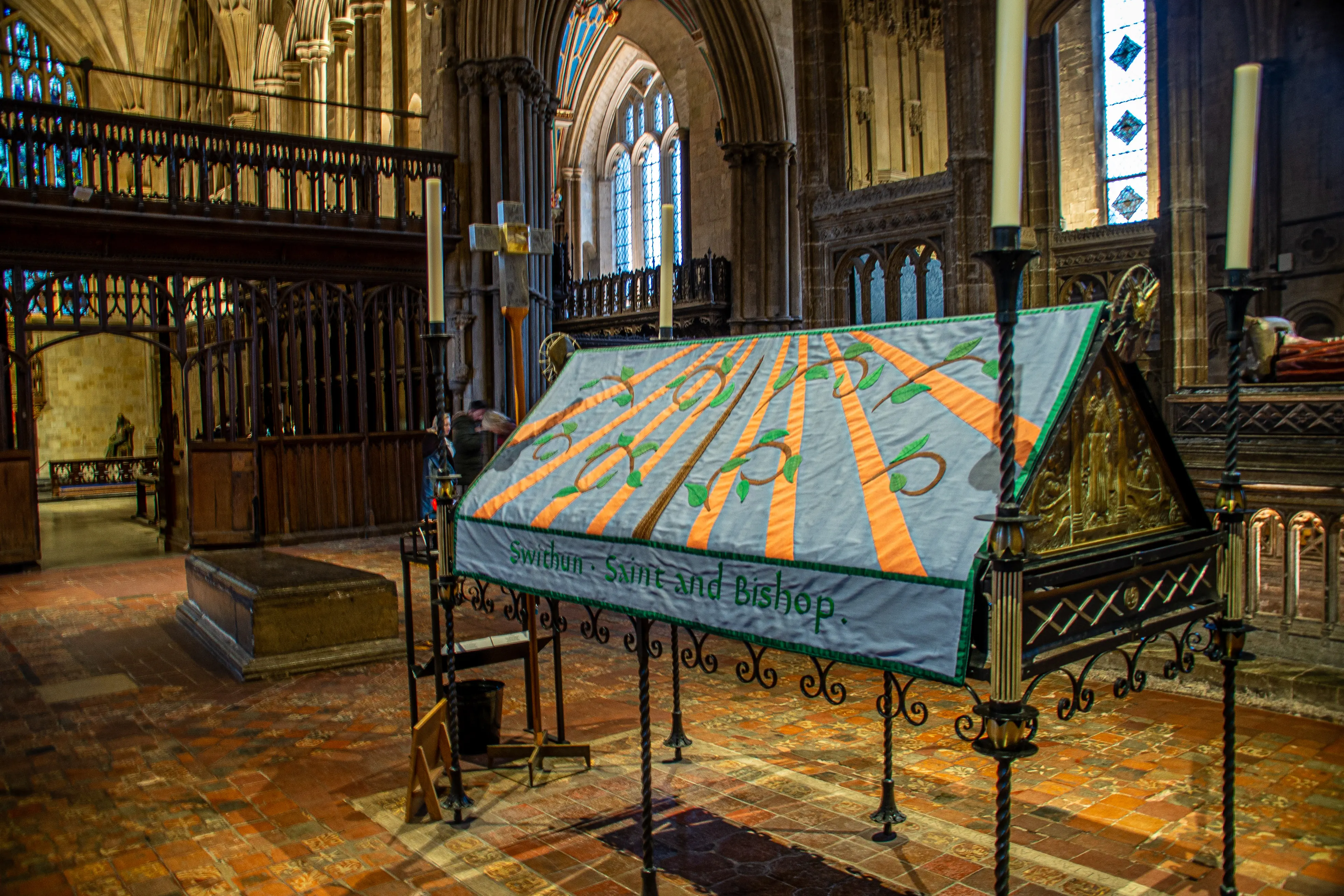 The image depicts the interior of a historic church or cathedral. The focal point is an ornate structure covered with a cloth that has the words 'Swithun, Saint and Bishop' embroidered on it. The setting includes stained glass windows, stone arches, and wooden furnishings, contributing to a serene and reverent atmosphere