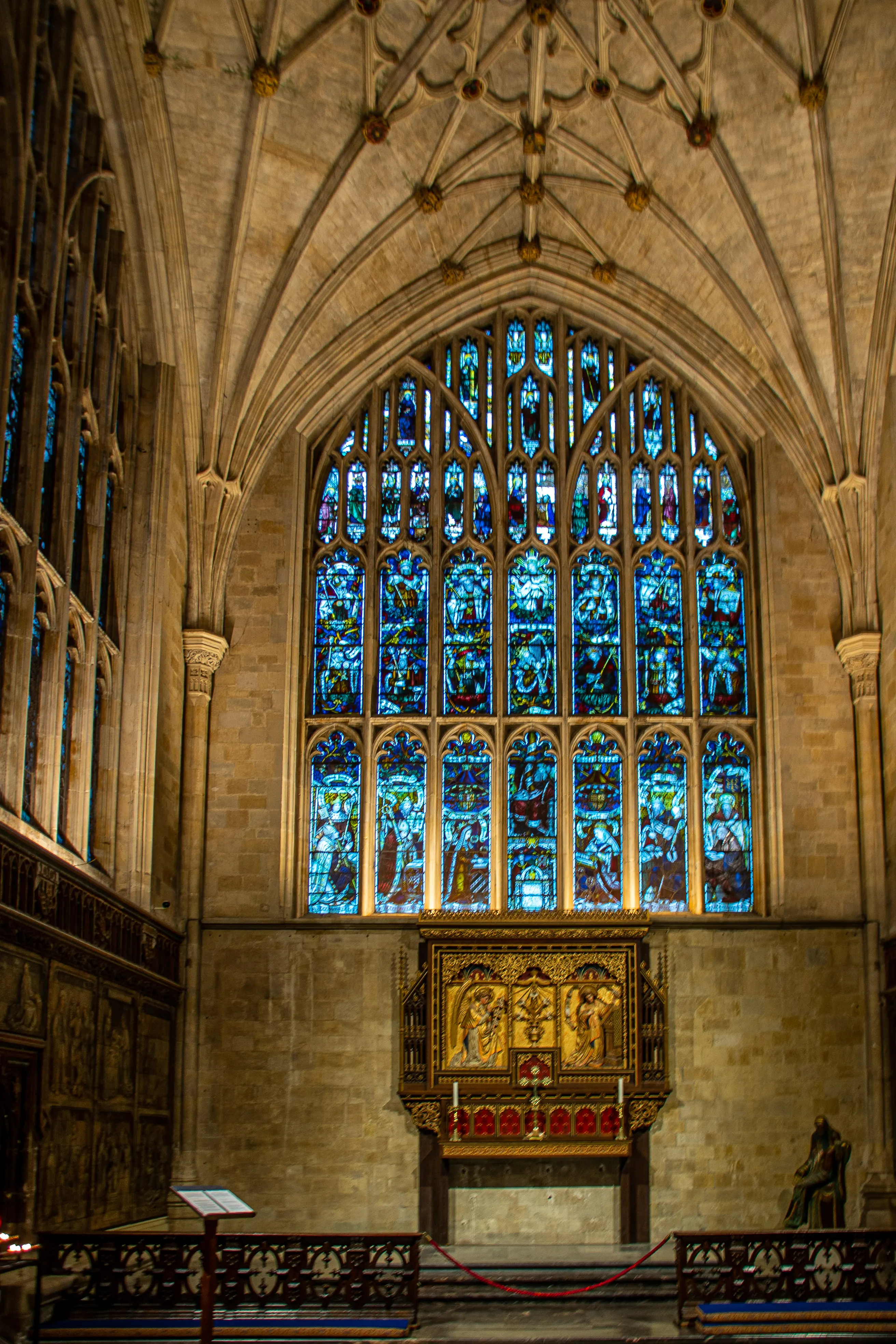 The image depicts the interior of a grand cathedral or church. It features a large, ornate stained glass window with intricate designs and vibrant colors. Below the window, there is an elaborately decorated altar or shrine with golden embellishments and religious iconography. The architectural elements include high vaulted ceilings with ribbed arches and detailed stonework. The overall ambiance is one of historical and religious significance.