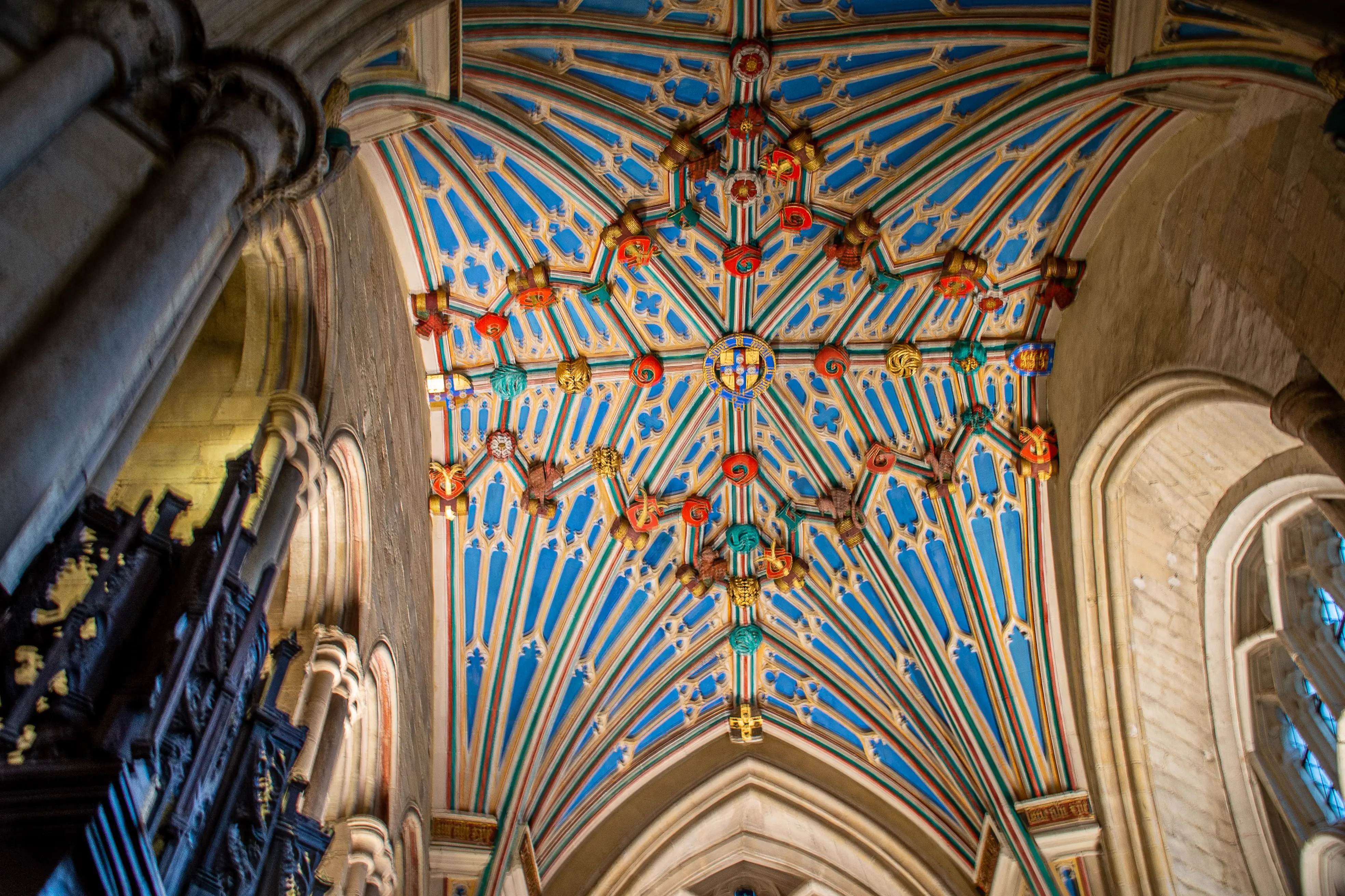 The image depicts an intricately designed ceiling of a historical building, likely a cathedral or church. The ceiling features a detailed pattern with vibrant colors, primarily blue, red, and gold. The architecture includes ribbed vaults and decorative elements such as shields and floral motifs. The surrounding stone columns and arches add to the grandeur and historical significance of the structure.